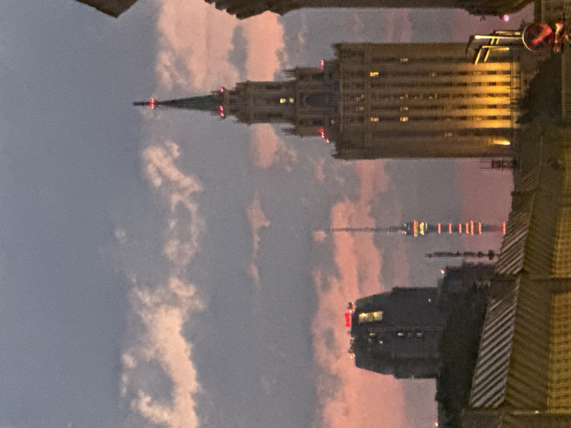 The image shows a twilight view of the Stalinist skyscrapers in Moscow, Russia. The primary subject is the Stalinist skyscraper, illuminated by warm lights. To its left, a darker, more modern building with red lights on its roof is visible. In the background, a radio tower with red and white lights stretches towards the sky. The sky is a dusky purple with streaks of pink from the setting sun, and clouds are scattered throughout. In the foreground, a darker structure, possibly a fence or a building facade with a patterned roof, is visible. The overall scene depicts an urban landscape at dusk.