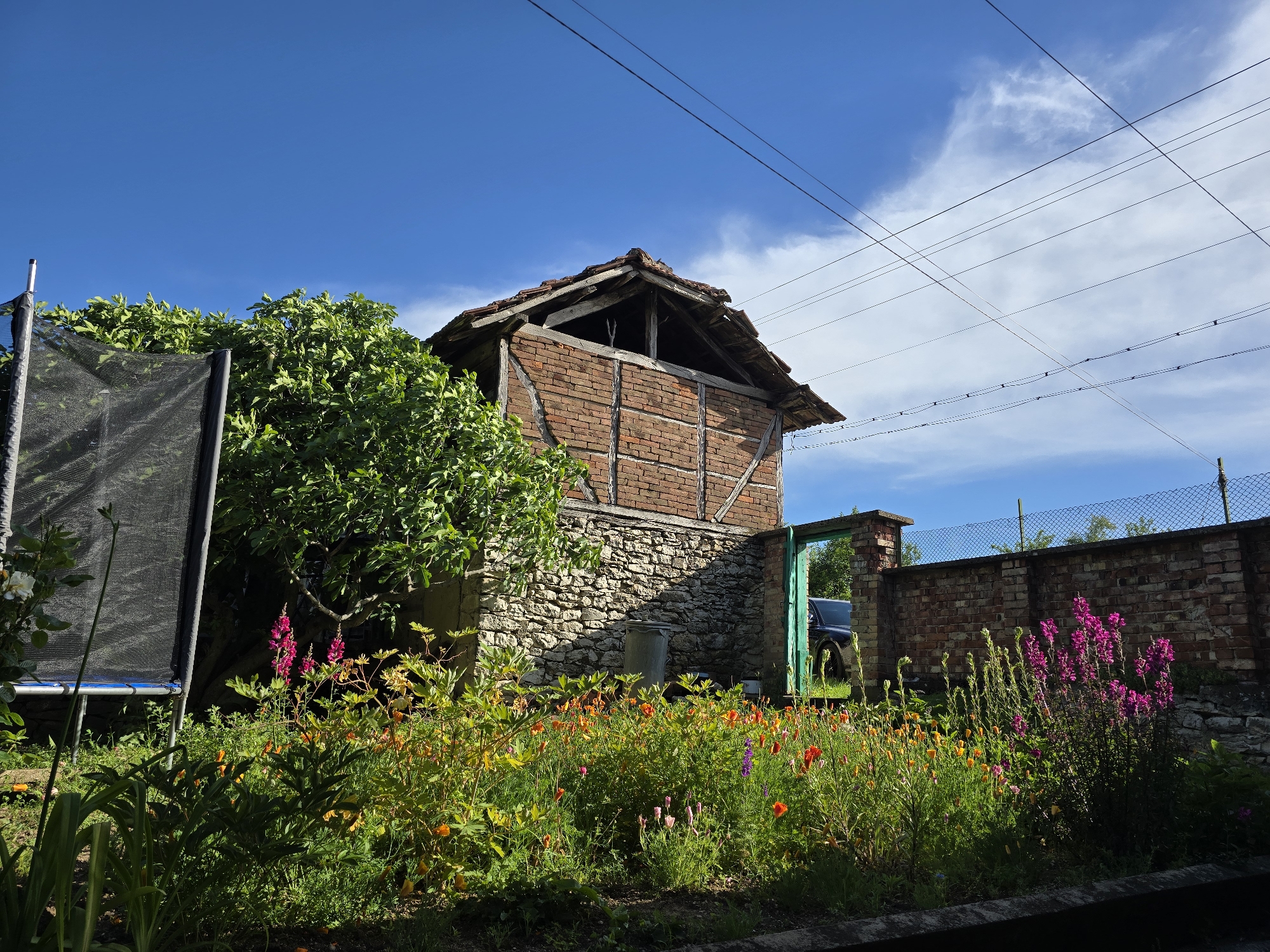 The image depicts a rural scene with an old stone and brick building dominating the view. The building has a stone foundation, a brick upper section, and a partially dilapidated roof. A green door is set within a brick and stone wall adjacent to the building, revealing a dark blue vehicle. In front of the building and wall is a flower garden, filled with a variety of colorful blooms. A trampoline with a black mesh safety net is visible to the left, partially obscured by a leafy tree. Overhead, power lines crisscross against a bright blue sky with scattered clouds. The location appears to be a backyard or garden setting in a rural or semi-rural area. The city and country are listed as "Unknown, Unknown."