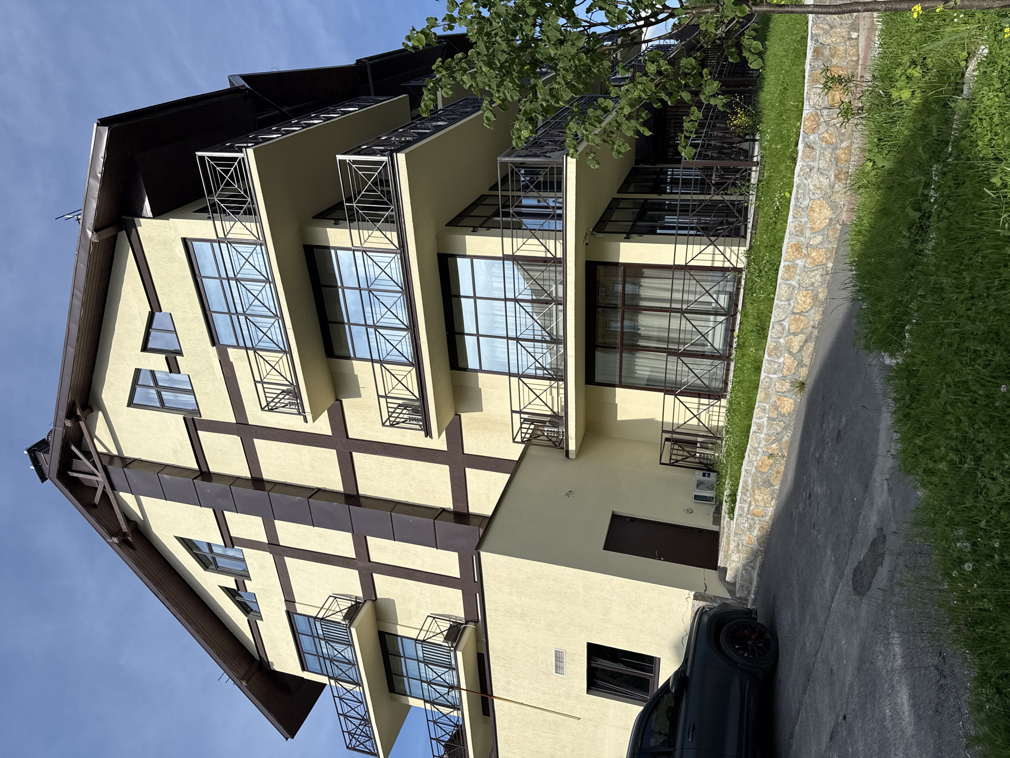 A low-angle exterior shot captures a modern, multi-story building with a light yellow facade, accented by dark brown vertical and horizontal beams in a grid pattern. The building features several balconies with black metal railings and large, dark-framed windows. The roof is dark brown and gabled, with exposed wooden structural elements visible under the eaves. A dark-colored SUV is parked on the asphalt in the foreground, to the left of the building. The building is situated on a slight incline, with a retaining wall made of light-colored stones separating a grassy area from the driveway. Green grass and a tree with lush foliage are visible on the right side of the image. The sky is a clear blue with a few wispy clouds. The scene appears to be in a suburban or resort setting, possibly in Syrostan, Russia, given the architectural style which is consistent with modern mountain resort accommodations. There are no people visible in the image.