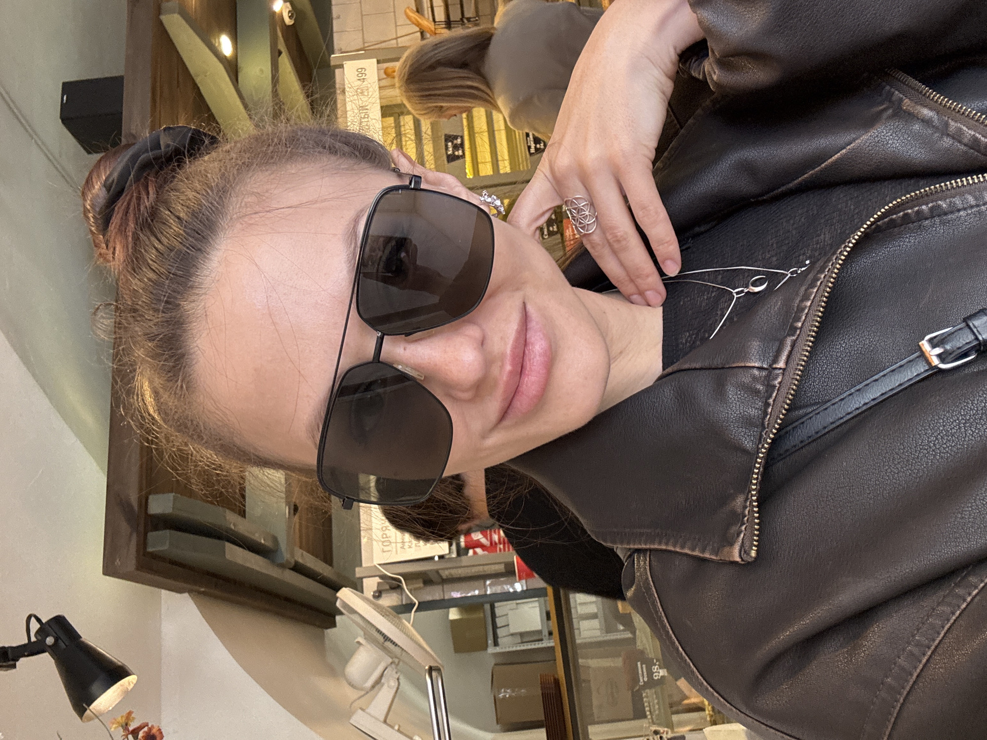 A person wearing dark sunglasses and a brown leather jacket poses indoors with their hand near a silver necklace. The location is suggested by text in Russian visible in the background, indicating a location in Saint Petersburg, Russia. The image is rotated counter-clockwise.