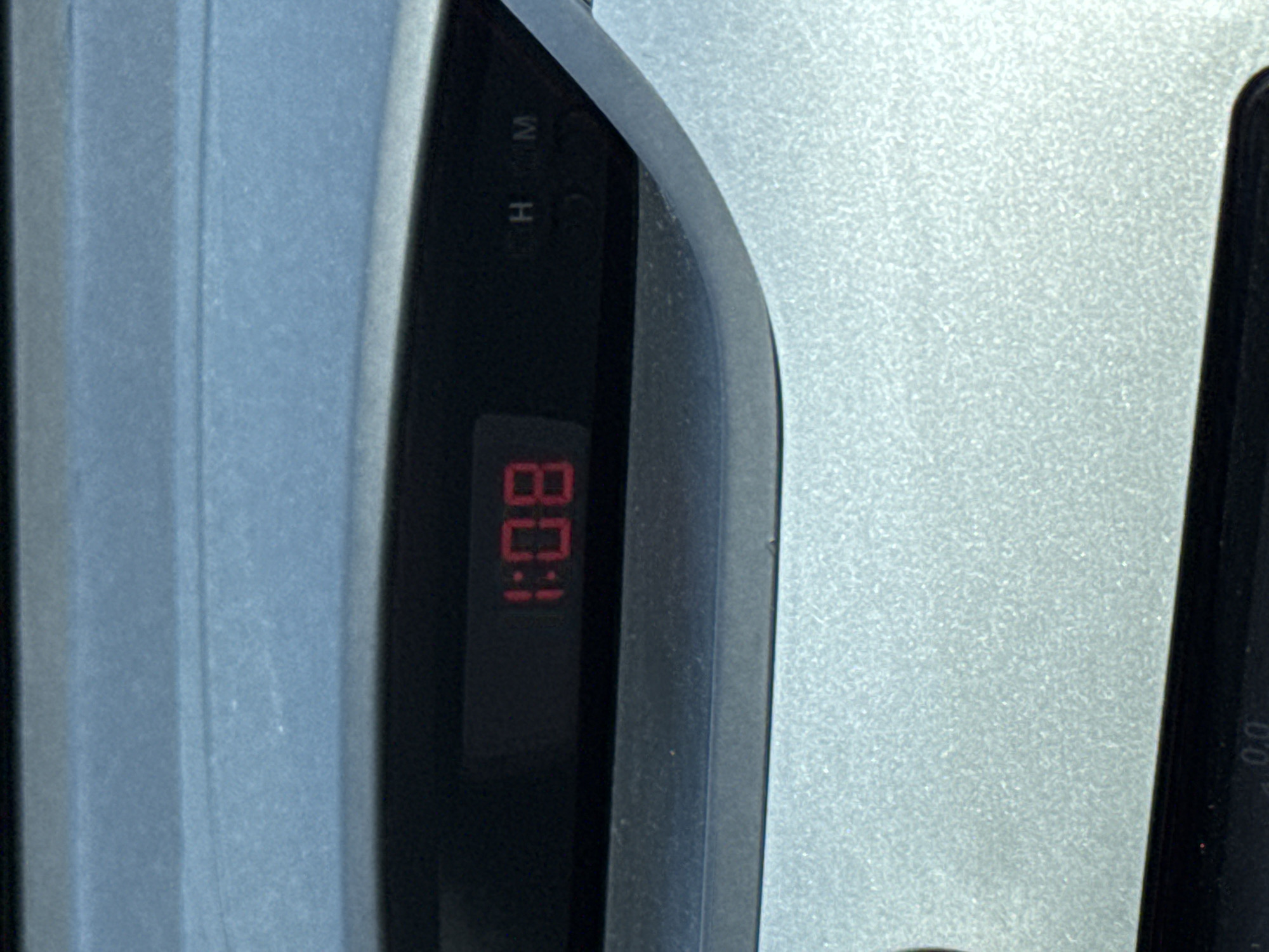 A close-up shot shows a digital clock displaying "1:08" in red numerals. Above the clock face are faint letters "H" and "M", possibly indicating hours and minutes. The clock is embedded in a dark gray or black panel. To the left of the panel is a light blue, textured surface, possibly a car interior. To the right, a silver or light gray textured surface is visible, potentially a dashboard component. The location in Bac Nha Trang Ward, Vietnam, is not evident from the image. The scene contains no people, actions, or interactions, but only the depiction of the clock integrated with the vehicle's interior.