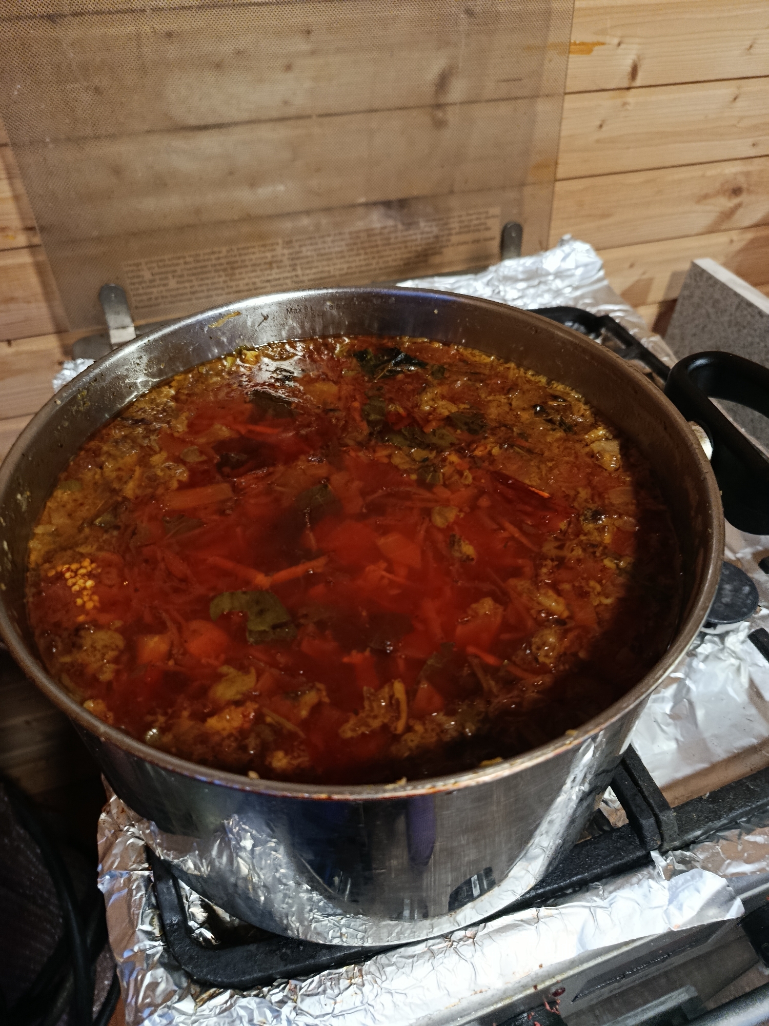 A large, metallic cooking pot, marked "MAX 8L" and "8.5" on its rim, is centered in the frame, containing a dense, red-orange soup or stew. The dish features visible ingredients including chopped root vegetables (e.g., carrots, beets), leafy greens, and a concentrated cluster of small, pale, granular ingredients (e.g., split peas, spices) on the left side. The liquid surface exhibits a slight oily sheen. The pot sits on a gas stovetop, which is covered with aluminum foil. A black handle is visible on the pot's right side. The background consists of light-toned wooden paneling, typical of an indoor kitchen environment in the city of Vereya, Russia. A semi-transparent, textured splash guard is positioned above the pot, revealing faint textual content through its surface. No individuals are present in the image.