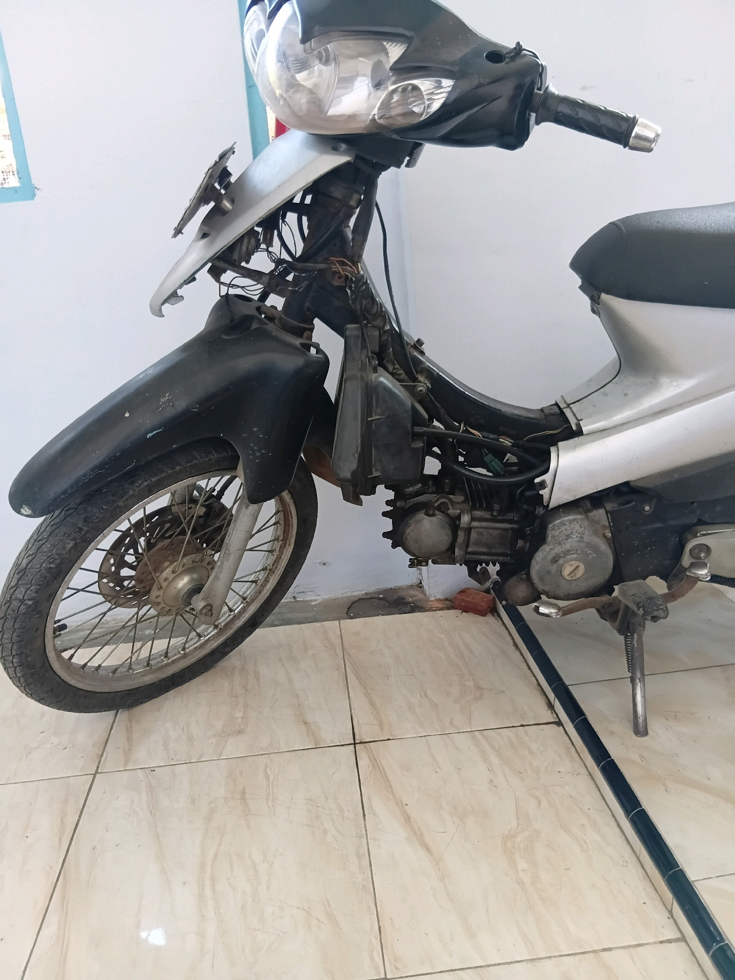 A partially disassembled silver and black motorcycle is parked on a tiled floor against a light blue wall. The motorcycle's front fender and some of its body panels are removed, revealing the engine and wiring. The motorcycle is leaning on its kickstand. A black handlebar with a silver grip is visible at the top of the image. The floor is covered with light beige tiles with a subtle marbling pattern, and a dark blue and silver tiled trim runs along the bottom right of the image. A red brick is on the floor near the kickstand. The scene appears to be indoors, possibly in a workshop or garage. The location is identified as Tanggul Wetan, Indonesia.