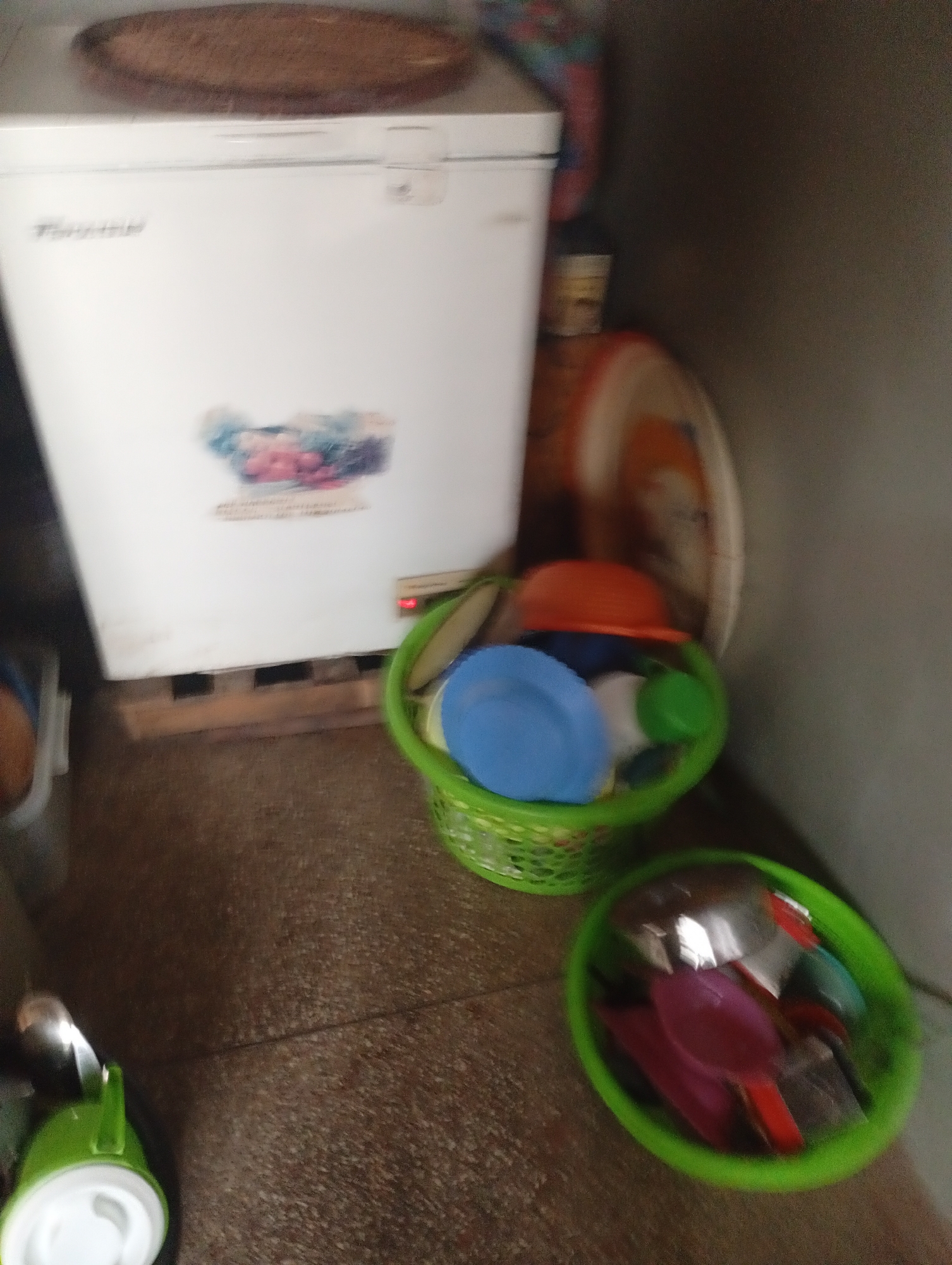 In a dimly lit room, likely a kitchen or storage area, a white chest freezer stands against a wall. A circular, brown object rests on top of the freezer. To the right of the freezer, a woven green basket is filled with various colorful plastic dishes and bowls, including blue and orange ones. Another similar green basket, positioned lower and to the right, contains a collection of kitchen utensils, such as spatulas and spoons, in shades of pink and red, alongside metallic items. In the foreground, a green pitcher with a white lid is visible, partially obscured by the frame. The floor appears to be tiled with a speckled brown pattern. The environment suggests a functional, albeit cluttered, domestic space.