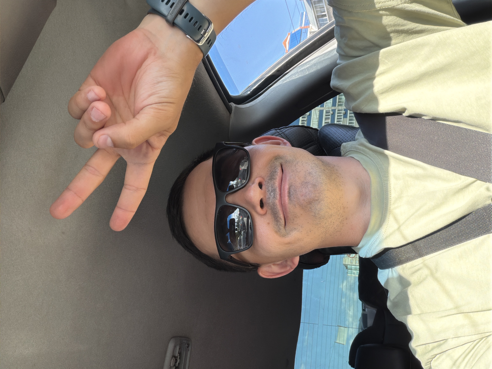 A man is in the front seat of a car. He wears sunglasses and a seatbelt, and is giving a peace sign with his left hand. The car's sunroof is open, revealing a blue sky. Buildings are visible through the sunroof. The reflection in the sunglasses shows buildings across the street, which are in Izhevsk, Russia.