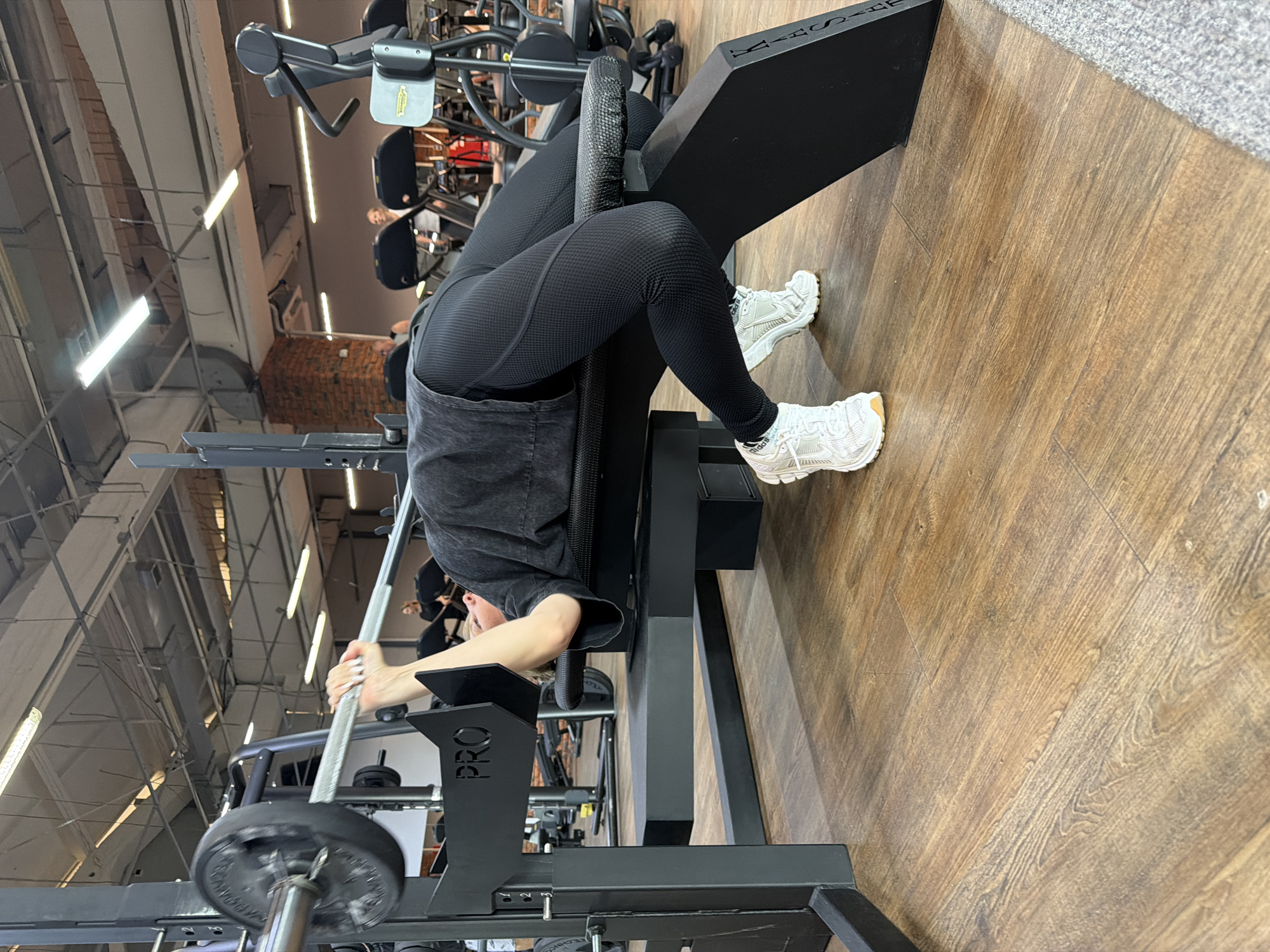 A woman is performing a barbell hip thrust exercise on a workout machine in a gym. The machine is black with the word "PRO" visible on one of the upright supports. The woman is wearing black leggings, a gray top, and white sneakers. She is lying on the padded seat of the machine with her shoulders supported, and her feet are on the floor. She is holding a barbell across her hips. The floor is made of wood. Gym equipment is in the background. The location is Chaikovsky, Russia.