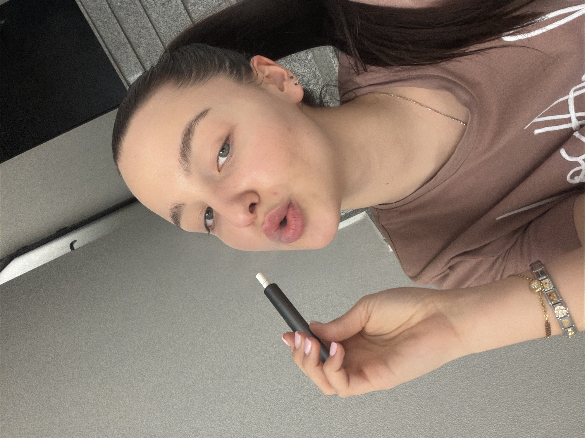 A young woman with fair skin and dark hair pulled back into a ponytail is holding a black electronic cigarette in her right hand. She is wearing a brown t-shirt with white designs and a gold chain necklace. The background appears to be a wall, partially gray and partially a textured material. The image is rotated counterclockwise. There is no information present that supports the claim that the image was taken in Sofia, Bulgaria.