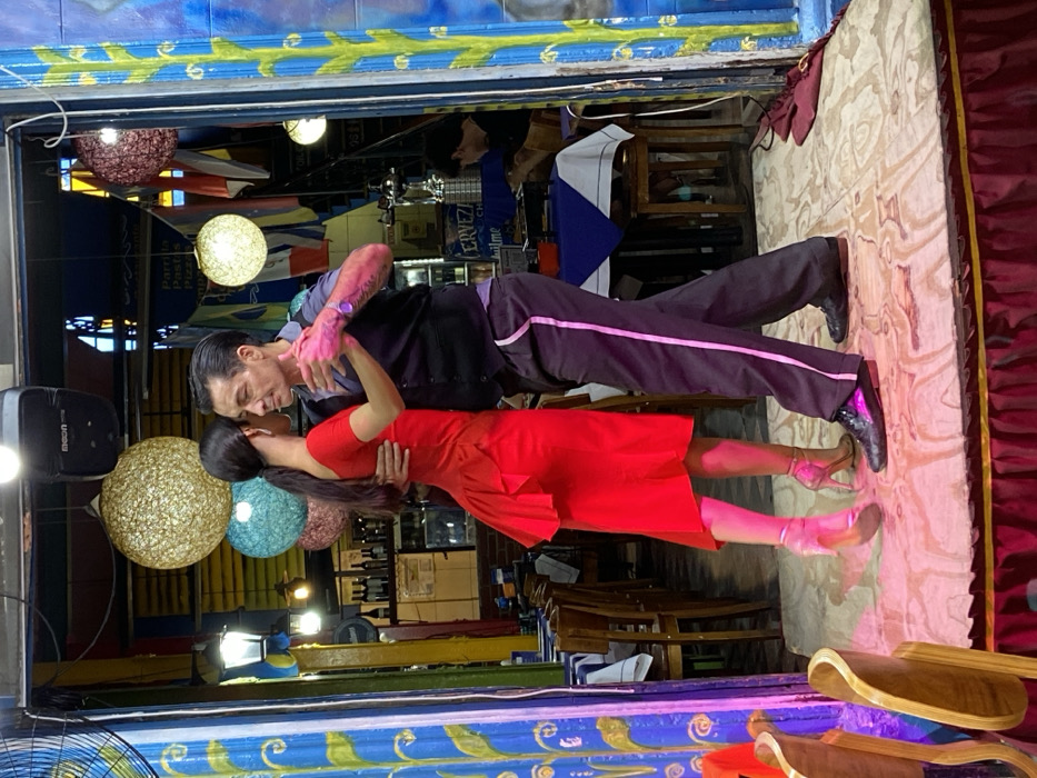 Inside a colorful building, a tango couple dances on a stage. The man, with dark hair and visible tattoos, wears a dark vest and pants with a pink stripe. The woman is in a red dress and matching heels. They are captured mid-dance in an embrace. Light fixtures hang near them. The background includes tables, chairs, flags, and a bar. The overall atmosphere is vibrant and lively.