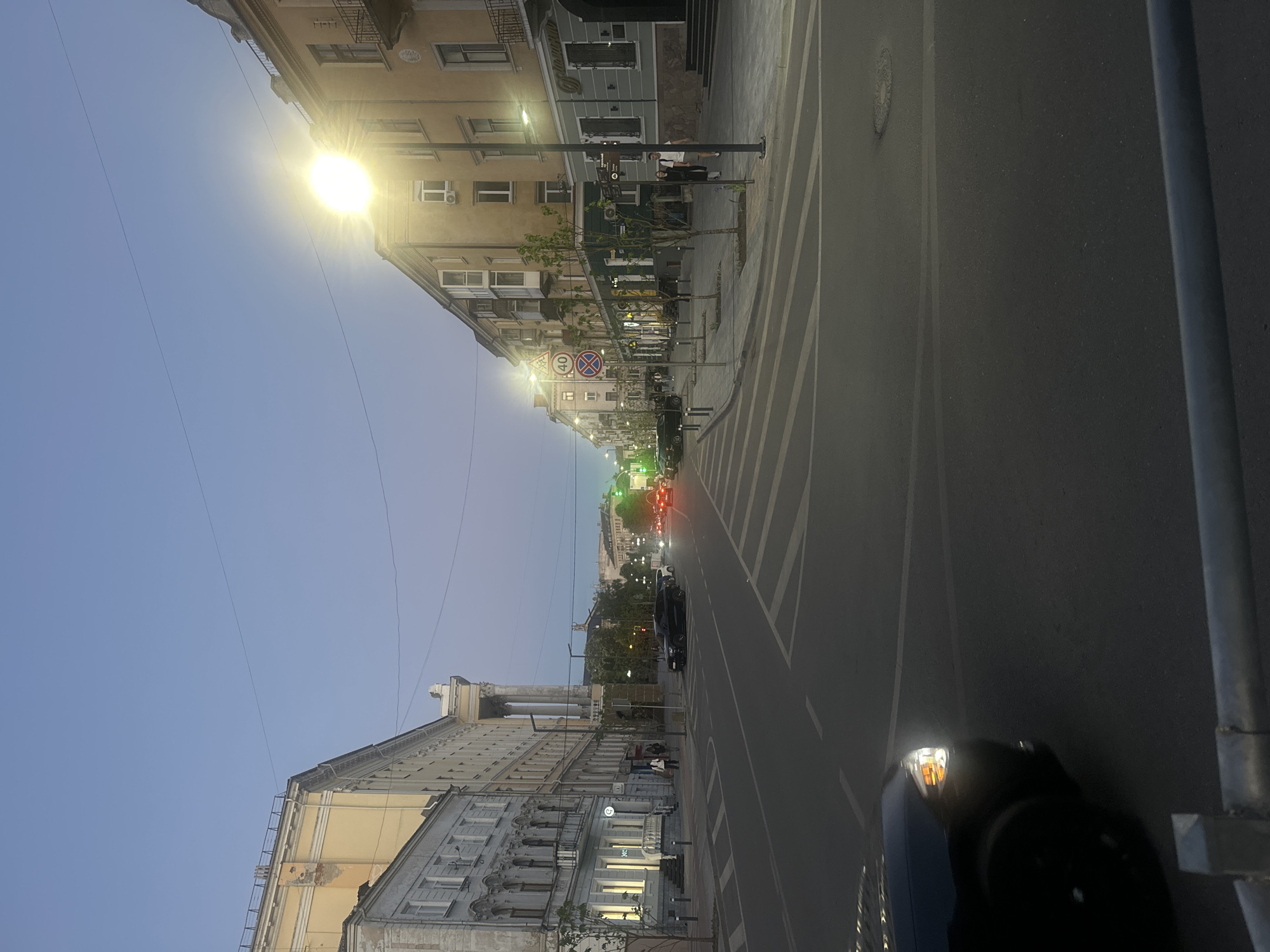 The image shows a street scene in Ternopil, Ukraine. The view looks down a street with buildings on either side. The buildings vary in color and architectural style. There are pedestrian crosswalks visible. Street lights are on. Cars are present on the street. The sky is a gradient from light blue near the horizon to a darker blue above. A traffic sign and other street signs are visible along the street. A large arch-like structure is visible further down the street. A vehicle part is visible in the foreground.