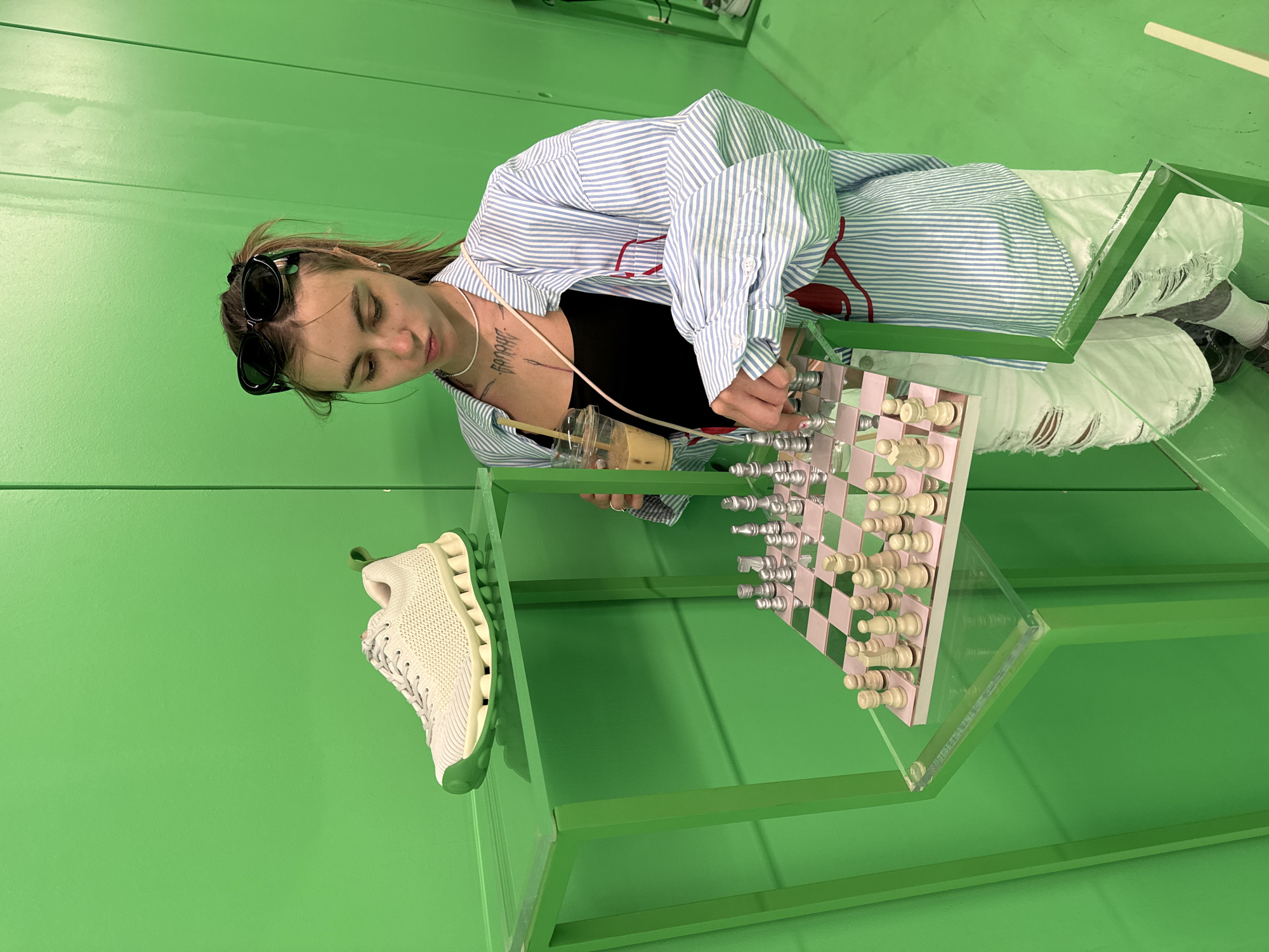 A young woman with sunglasses perched on her head and her hair pulled back in a ponytail stands in front of a bright green wall. She is wearing a blue and white striped open button-down shirt over a black tank top and distressed white jeans. She holds a to-go cup with a beverage in her left hand. In front of her, on a clear acrylic stand with green metal legs, is a chessboard with both light and dark wood pieces, and a white sneaker is displayed on a shelf above it. Her right hand is positioned over the chessboard, as if she is about to move a piece. The setting appears to be an indoor exhibition or retail space, likely in Tbilisi, Georgia, given the green backdrop often used in such environments.