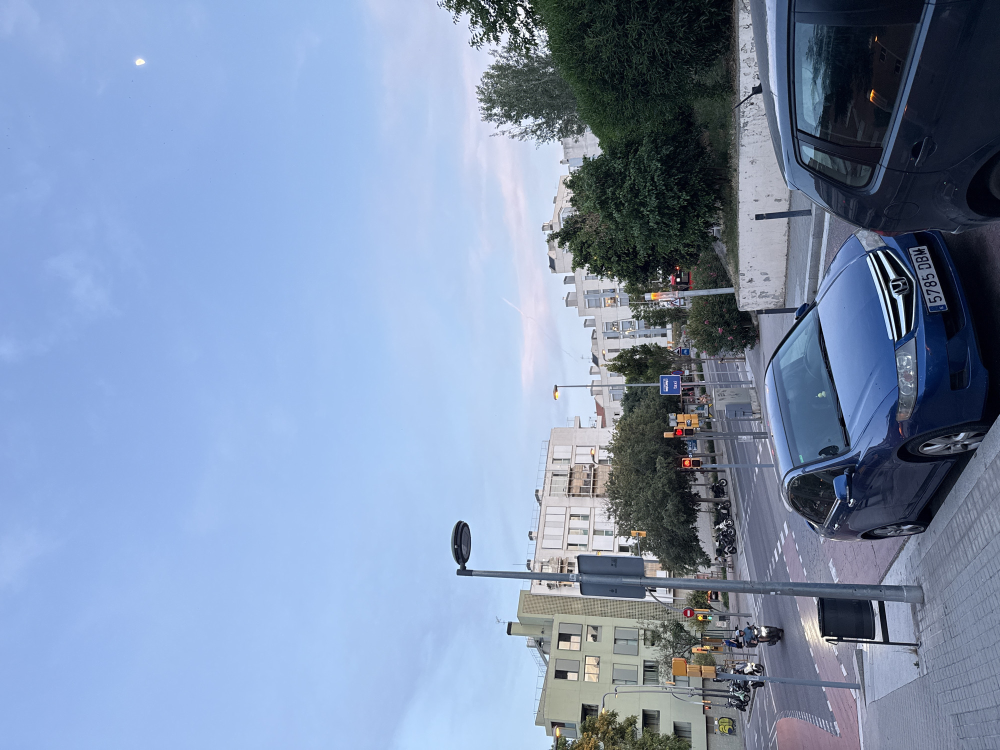 This image captures a street scene in Barcelona, Spain, at dusk. The sky is a gradient of pale blue and pink, with a sliver of the moon visible in the upper right corner. In the foreground, a blue sedan is parked on the right, and its license plate reads "5785 DBM". To its left, another dark-colored car is partially visible. A pedestrian sidewalk runs along the street, made of gray pavers.
The street itself is a two-lane road with marked lanes. Several motorcycles are seen riding on the road. Traffic lights are visible, some showing red. Buildings line the street, characterized by modern architecture with white and light green facades and numerous windows. Trees and green bushes are interspersed along the sidewalk and in front of the buildings, adding a touch of nature to the urban environment. Streetlights are also present. One prominent streetlight stands tall on the left side of the image. Signs are posted along the street, including a blue sign with "TAXI" written on it. The overall impression is a quiet evening in a well-developed urban area.