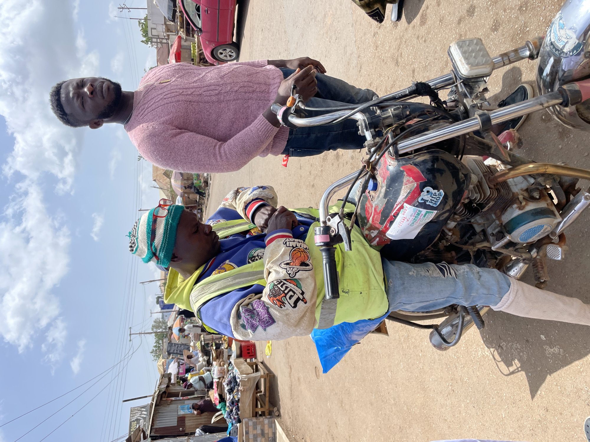 Two men stand next to a motorcycle on a paved surface. One man is seated on the motorcycle, wearing a yellow safety vest, a jacket with patches, jeans with a tear, and a hat. He also appears to have a bandage on his leg. The other man stands beside the motorcycle, wearing a pink sweater and jeans. Behind them, there is a bustling outdoor market scene with stalls and people, indicating a commercial area. In the background, buildings and a red car are visible. The environment appears to be a street in a city market in Gura Top, Nigeria.