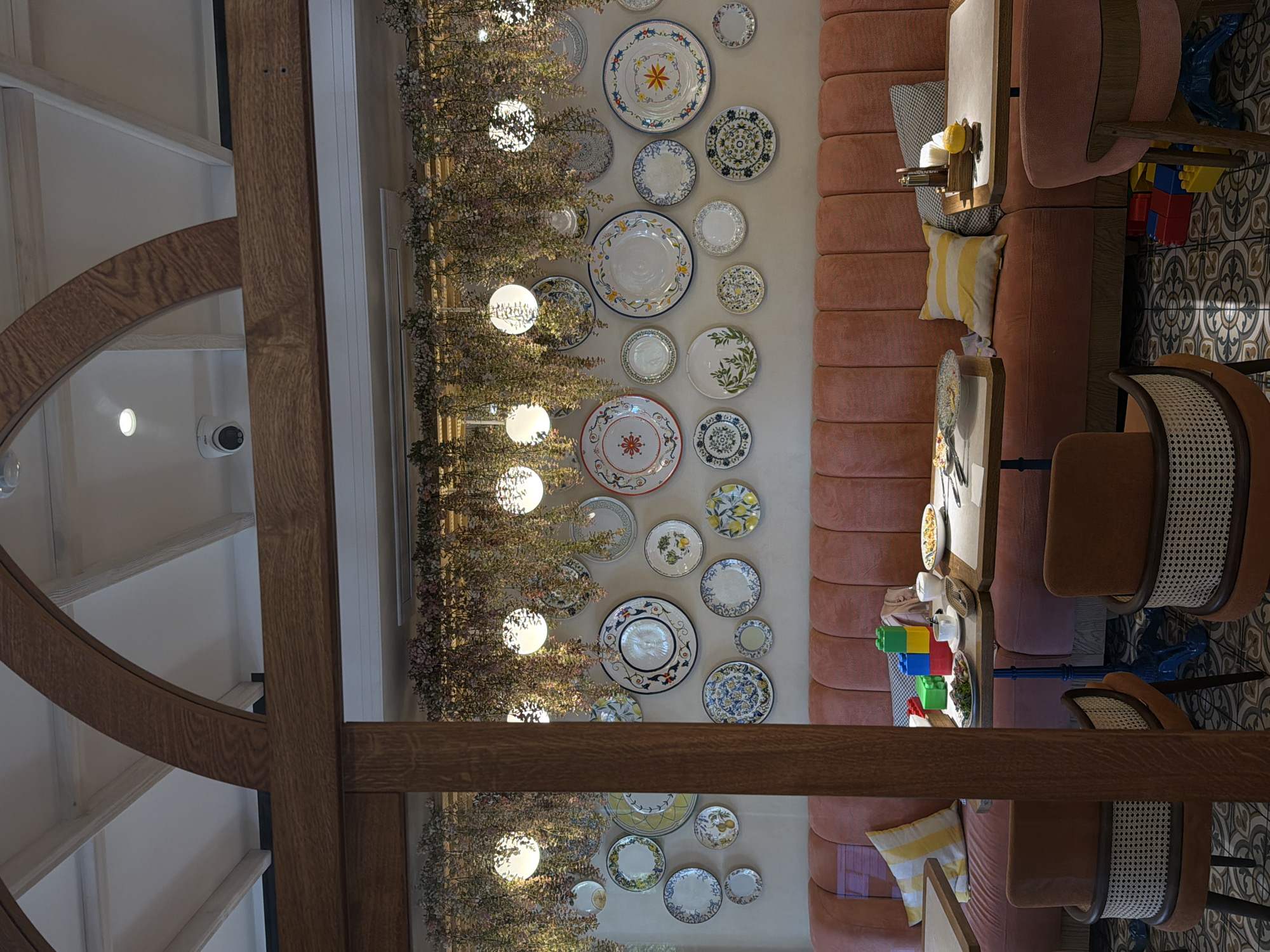 The image depicts the interior of a restaurant. The walls are adorned with a collection of decorative plates of various sizes and designs. Hanging plants and spherical lights are suspended from a wooden structure above the plates. A long, coral-colored banquette runs along the wall, complemented by wooden chairs with woven seats. On the tables, there are plates of food, cutlery, a coffee cup, building blocks, and a lemon. The flooring features a patterned tile design. The restaurant appears to be located in Volgograd, Russia, though no specific street address or building name is visible. There are no people clearly visible in the image.