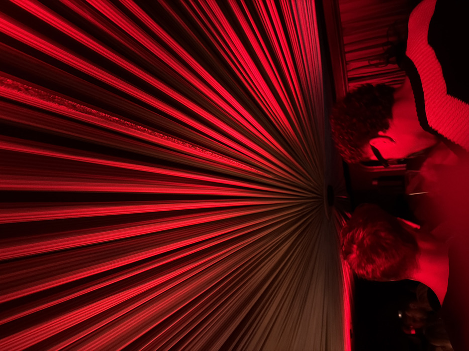The image depicts an indoor scene within a venue in Paris, France, illuminated predominantly by a strong red light. The upper portion of the frame is dominated by a ceiling structure featuring a radial fan pattern of draped or pleated fabric, with distinct red lines radiating outwards from a central point, against darker, shadowed channels. In the mid-foreground, two individuals are visible from behind, immersed in the red lighting. The person on the left has short, light-colored hair and is wearing a dark, possibly sleeveless top. The individual on the right has dark, curly hair, wears dark glasses, and is dressed in a dark garment with a distinct white or light-colored striped pattern across the shoulder and upper back. Both appear to be observing a point beyond the frame, facing away from the viewer. Indistinct, blurred figures are visible in the darker background to the left, suggesting the presence of a crowd.