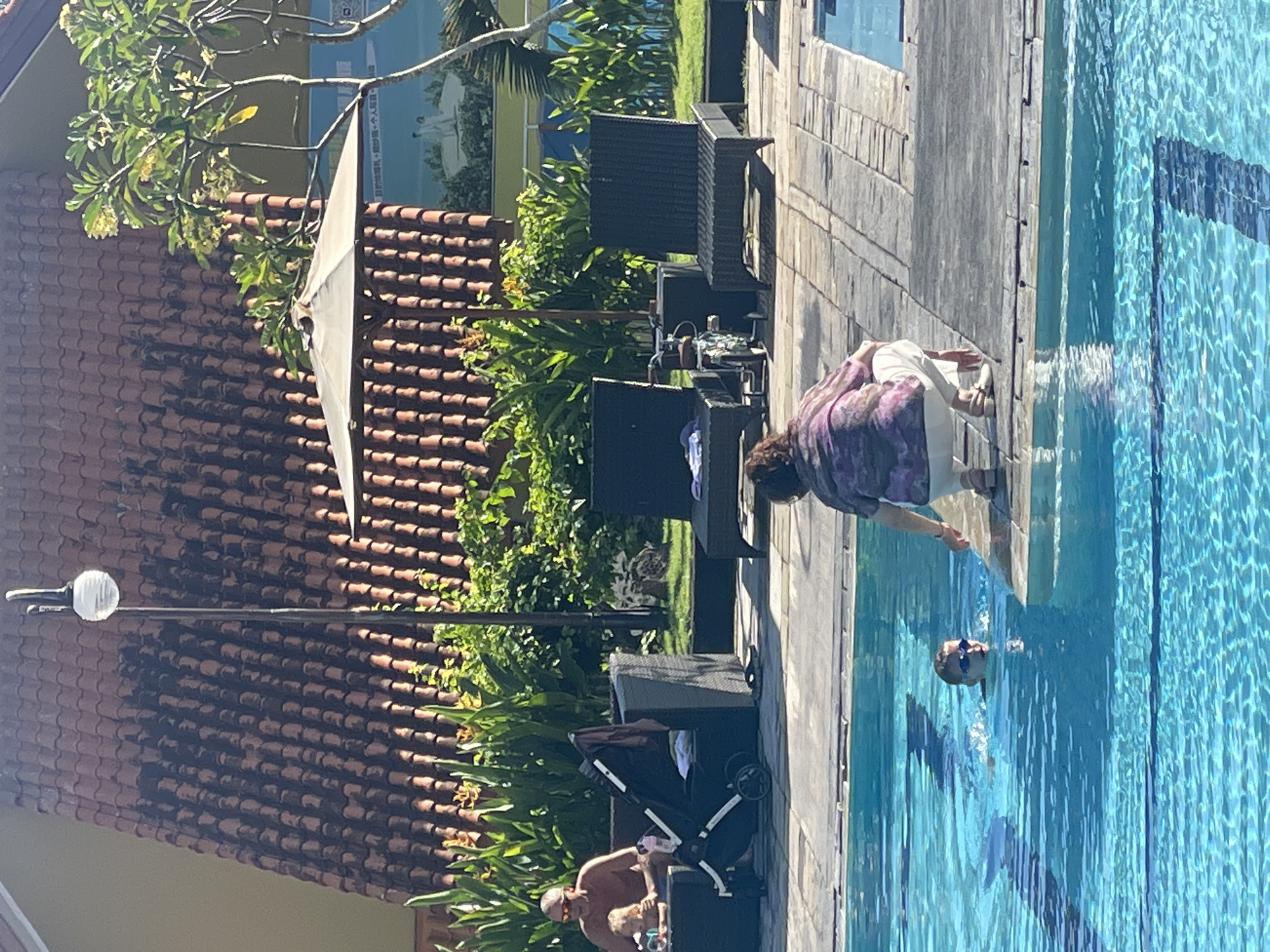 The image depicts a sunny outdoor scene at what appears to be a resort or hotel pool area in Kuta Selatan, Indonesia. In the foreground, a young boy wearing goggles is swimming in the bright blue water of the pool. A woman, wearing a patterned top and white pants, is kneeling at the edge of the pool, seemingly interacting with the boy. To the right, a portion of another person is visible.
In the background, beyond the pool deck, lounge chairs and a stroller are arranged near a building with a terracotta-tiled roof. Lush green foliage and trees are visible, along with outdoor lighting fixtures and a large umbrella. A mural or painted scene is visible on the wall of the building in the distance, depicting two figures in white on a grassy knoll. The overall atmosphere suggests a leisurely day at a tropical vacation spot.