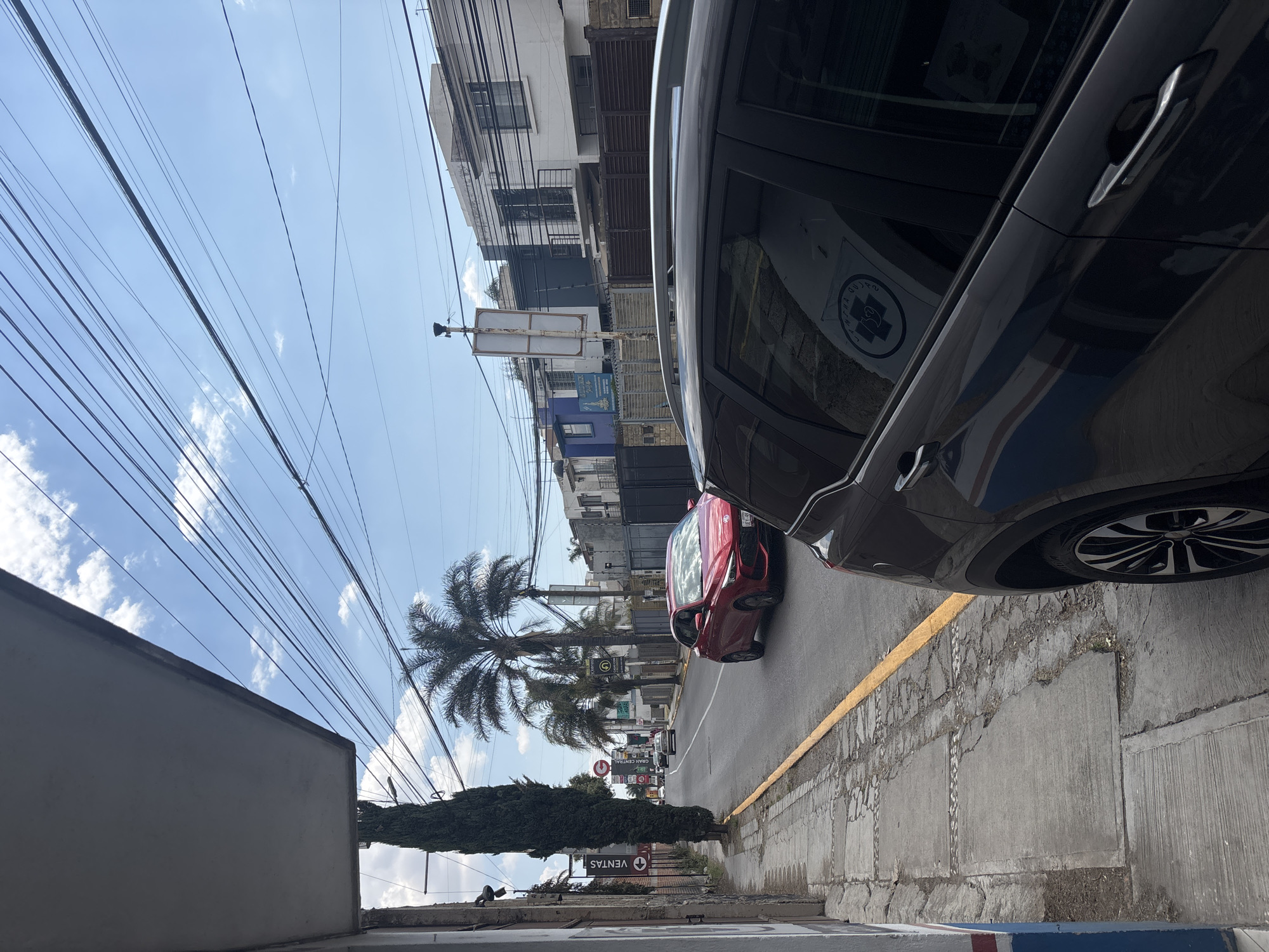 The image shows a street in San Juan Cuautlancingo, Mexico, taken from a sidewalk with a building on the left side. Overhead, power lines stretch across a partly cloudy sky. A tall, narrow tree and a “VENTAS” sign are visible in the foreground. Further down the street, palm trees line the road. A red car is driving on the road, and a larger black car is parked on the right side of the frame, partially obscuring the view. Buildings line the street on both sides.