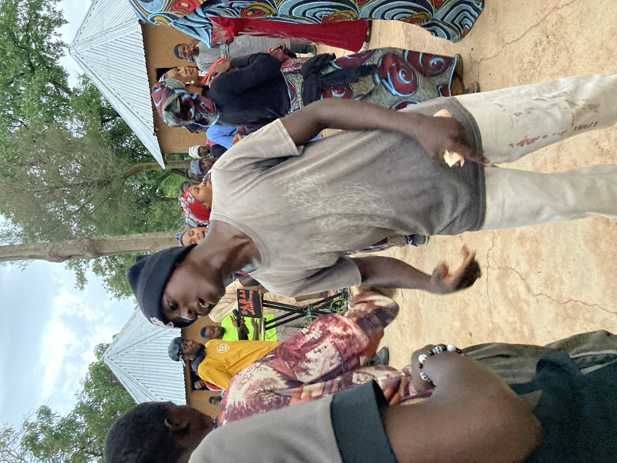 The image depicts an outdoor gathering of people, possibly at a community event or celebration, near structures with corrugated metal roofs, consistent with rural or semi-urban settings like those found in Jos, Nigeria. The ground appears to be a light-colored, dry earth or sand.
In the foreground and center, a young Black male wearing a black beanie, a grey t-shirt, and light-colored pants is the most prominent figure. His mouth is open as if speaking or singing, and his right hand is extended slightly outward, holding a small, light-colored object. His pants have red smudges and dirt marks. To his left, another Black male is partially visible, wearing a patterned garment over a grey shirt, and facing away from the camera.
In the background, several other individuals are present. A Black male in a bright yellow shirt with a logo and a black cap is standing near a DJ setup. The DJ setup includes a table with a laptop or keyboard and a speaker. Other people, some wearing headscarves in vibrant colors and patterns, are visible around the DJ area and further in the background, suggesting a lively atmosphere. The sky is overcast, indicating diffused lighting. The overall scene captures a moment of social interaction and potential musical entertainment.