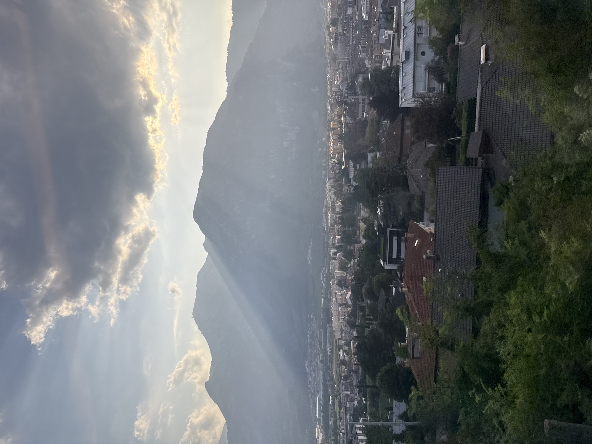 This image captures a hazy, sunlit mountain range behind the city of Trento, Italy. The sun's rays pierce through dramatic, grey clouds, casting beams of light onto the mountains and the city below. In the foreground, a cluster of houses with red and grey tiled roofs are partially obscured by lush green trees and foliage. Further into the distance, the urban landscape of Trento unfolds, with numerous buildings of varying sizes and architectural styles. The overall atmosphere is serene, with the natural grandeur of the mountains meeting the man-made environment of the city.