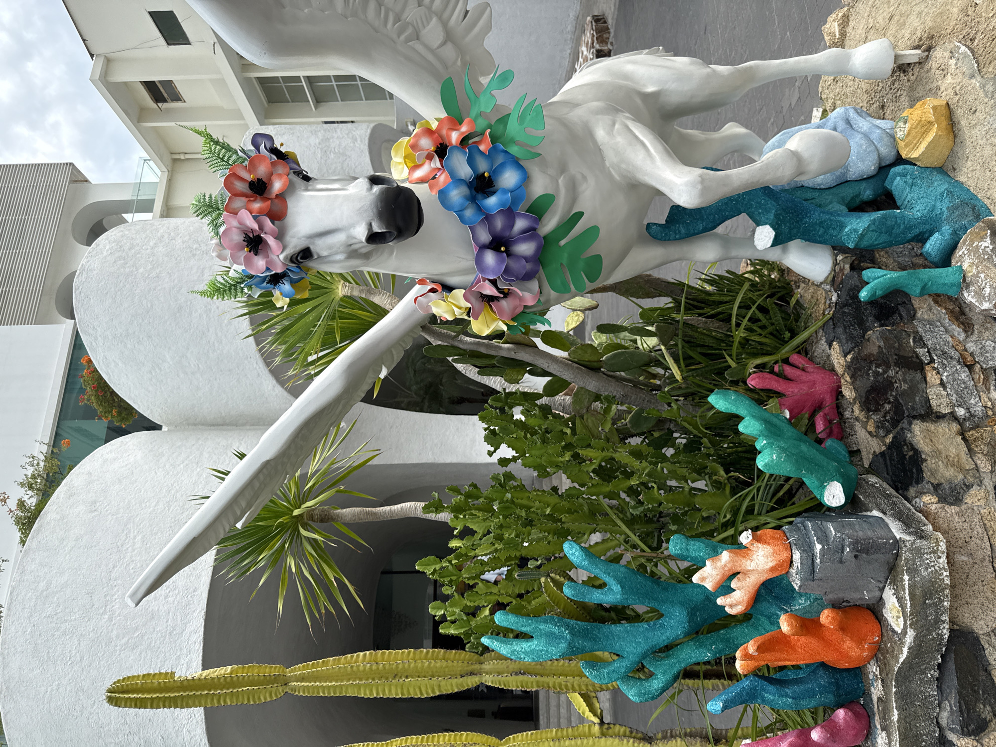 The image depicts a white statue of a Pegasus adorned with a floral crown and a garland of colorful flowers around its neck. The statue is positioned in front of a white building with curved architectural elements, possibly a hotel or resort in Nha Trang, Vietnam. Several cactus plants of varying sizes and a palm tree are visible behind the statue. In the foreground, there is a decorative arrangement of brightly colored, coral-like sculptures and rocks, creating an underwater or marine theme. The sky is overcast.