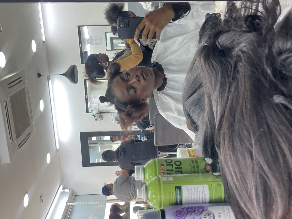 The scene depicts the interior of a hair salon in Paris, France. A dark-skinned female individual, wearing a white protective cape, is seated and captured in a mirror reflection taking a selfie with a black smartphone held in her left hand. Her natural hair is sectioned into multiple small, reddish-brown twists on her scalp, indicating preparation for styling. Dark lipstick is visible on her lips.
In the mirrored background, a female hairdresser with a high ponytail and large gold hoop earrings is visible to the right, wearing a light brown top and holding a yellow object, appearing to work on a client's hair. To the far left of the reflection, another person in a grey shirt and blue cap is seen from behind near shelves displaying several wigs on mannequin heads. Another client's hair, styled in a high bun, is partially visible to the right of the main subject's head.
The immediate foreground of the reflection shows dark hair extensions resting on a surface, alongside various hair product containers, including two green "ORS OLIVE OIL" spray cans and a purple "Soft as can be" mousse can. The salon environment features white walls, multiple large mirrors, recessed ceiling lights, a long fluorescent light strip, a black pendant light, and a ceiling-mounted air conditioning unit.