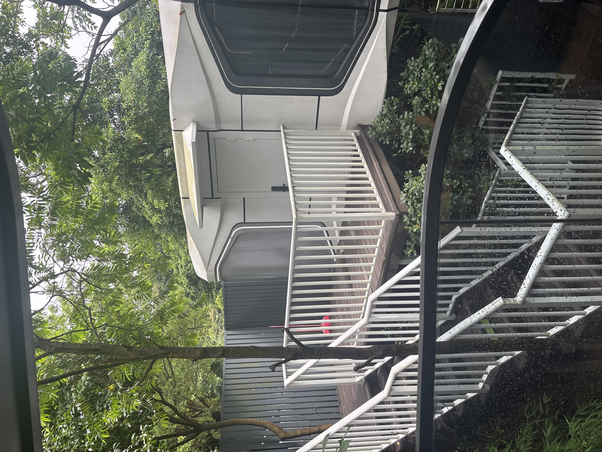 The image shows an outdoor scene with a modern, modular building that appears to be a pod-like structure. The building is white with dark trim around large, tinted windows. It has a single door with a handle and a small awning overhead. A wooden deck with white railings surrounds the entrance to the pod.
A set of white-railed stairs leads up to the deck. These stairs are set against a backdrop of lush green trees and foliage, suggesting a natural or semi-natural environment. In the distance, a grey metal fence is visible. A red broom is propped up against the railing on the deck. The stairs in the foreground also have white railings and appear to be wet, possibly from rain. The overall impression is that of a unique accommodation or retreat located in a verdant setting. The presence of trees, dense greenery, and the style of the building suggest it could be located in a park, a resort, or a residential area with extensive landscaping.