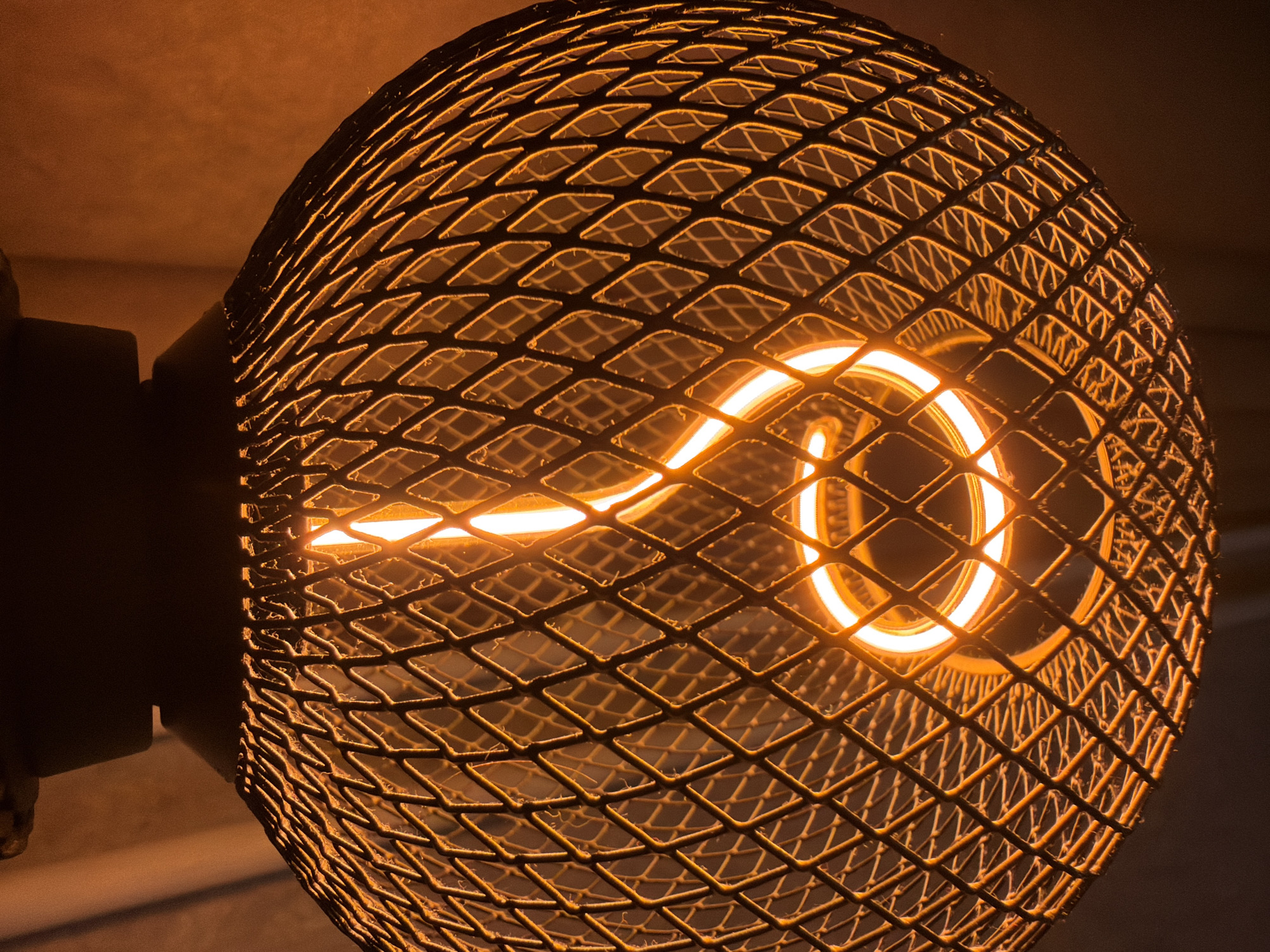 A close-up, low-angle shot captures a decorative light fixture, likely indoors during the evening or night. The central subject is an Edison-style LED light bulb, emitting a warm, bright orange-yellow glow. Its prominent feature is a visible, squiggly filament arranged in an artistic, curvilinear shape that resembles a stylized 'S' or spiral.
The light bulb is encased within a spherical or egg-shaped lampshade made of dark metal. This lampshade features a distinct diamond or rhomboid mesh pattern. The edges of the metal structure catch and reflect the warm light from the bulb, creating a luminous outline against the darker interior and exterior. The texture of the mesh appears slightly rough, contributing to an industrial yet elegant aesthetic.
The background is dark, soft, and out of focus, providing minimal environmental detail but emphasizing the light source. Hints of darker, structural elements are visible on the left side, suggesting the mounting point of the fixture. The overall scene is dominated by warm orange and dark tones, evoking a cozy or atmospheric ambiance. The image provides no visible text. This fixture is located in Sofia, Bulgaria.