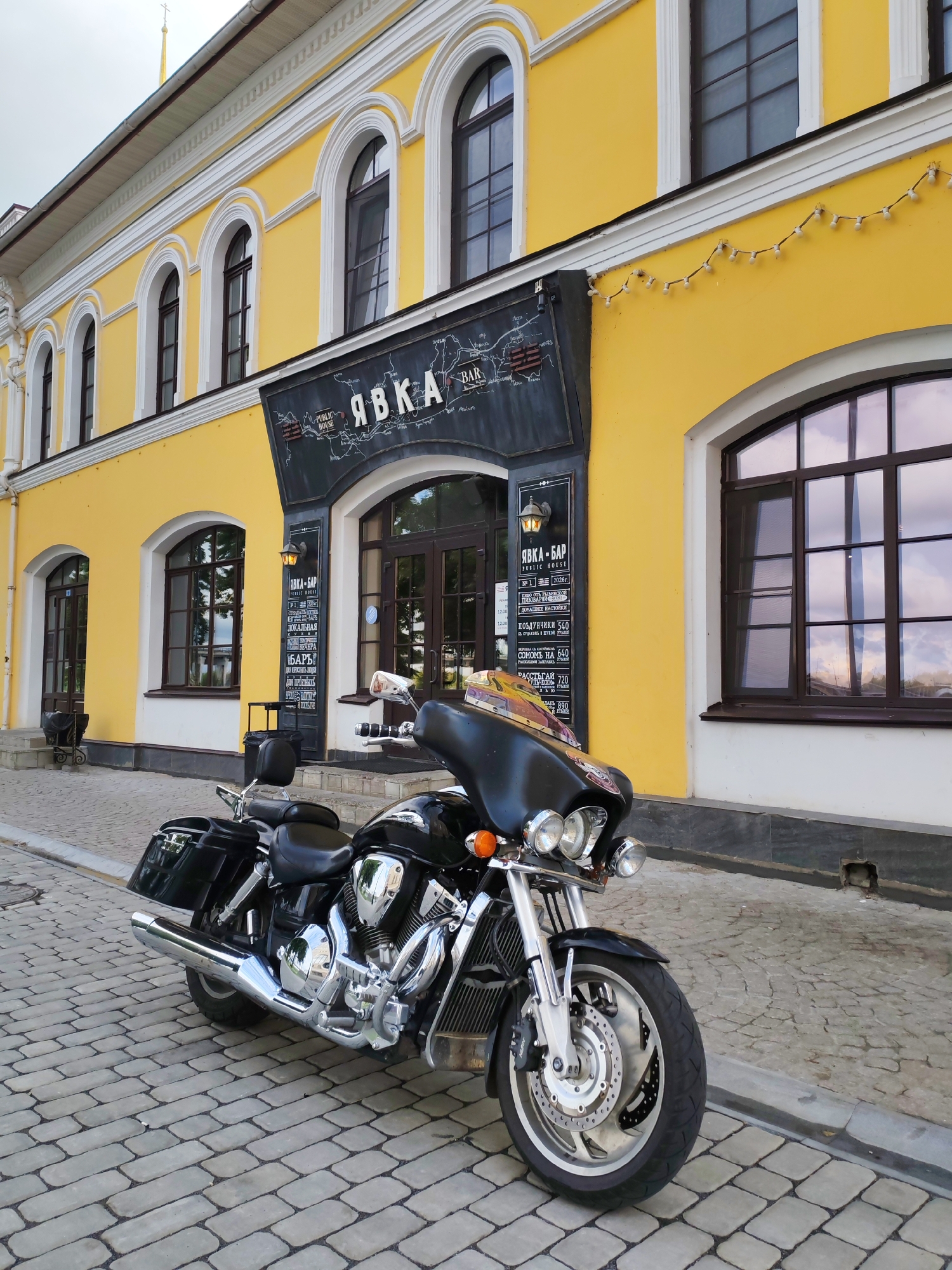 A black motorcycle is parked on a cobblestone street in front of a bright yellow building with arched windows. The building has a sign that reads "ЯВКА-БАР PUBLIC HOUSE." The motorcycle is the main subject, detailed with chrome accents and a black leather seat. The environment is an urban setting, with the building appearing to be a commercial establishment. The time of day appears to be daytime, with overcast skies. There are no people visible in the frame. The motorcycle is positioned diagonally, with its front wheel closer to the viewer. The cobblestone street has a distinct pattern. The building exhibits architectural details consistent with older European construction.