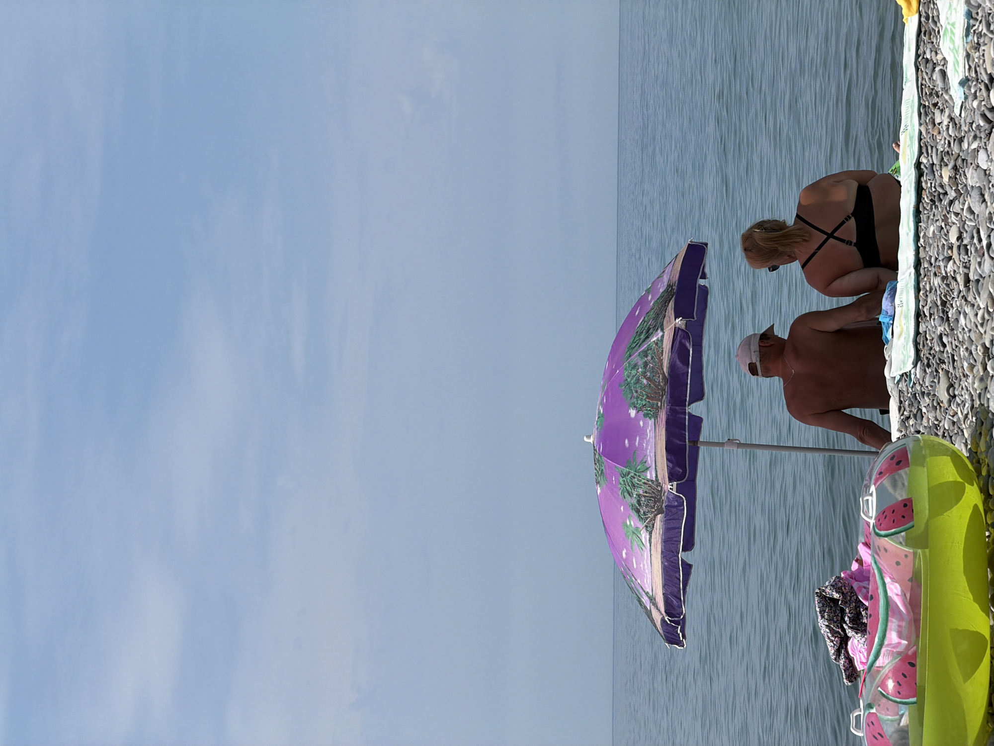 This is a full, vertical shot of a beach in Sochi, Russia, on a clear, sunny day. The camera is positioned low, facing the sea. In the foreground, a large, round, inflatable tube decorated with watermelon slices sits on the pebble beach. Behind it, a couple is seated on a towel, facing away from the camera and towards the calm, blue sea. The man, wearing a white baseball cap, has his back to the viewer, and the woman is wearing sunglasses and a black bikini top. They are positioned under a large, purple beach umbrella adorned with a tropical scene of palm trees. The umbrella's pole is visible, planted in the sand. The sea stretches out to the horizon, meeting a pale blue sky with a few wispy white clouds. The overall impression is one of a peaceful, summer day at the beach.