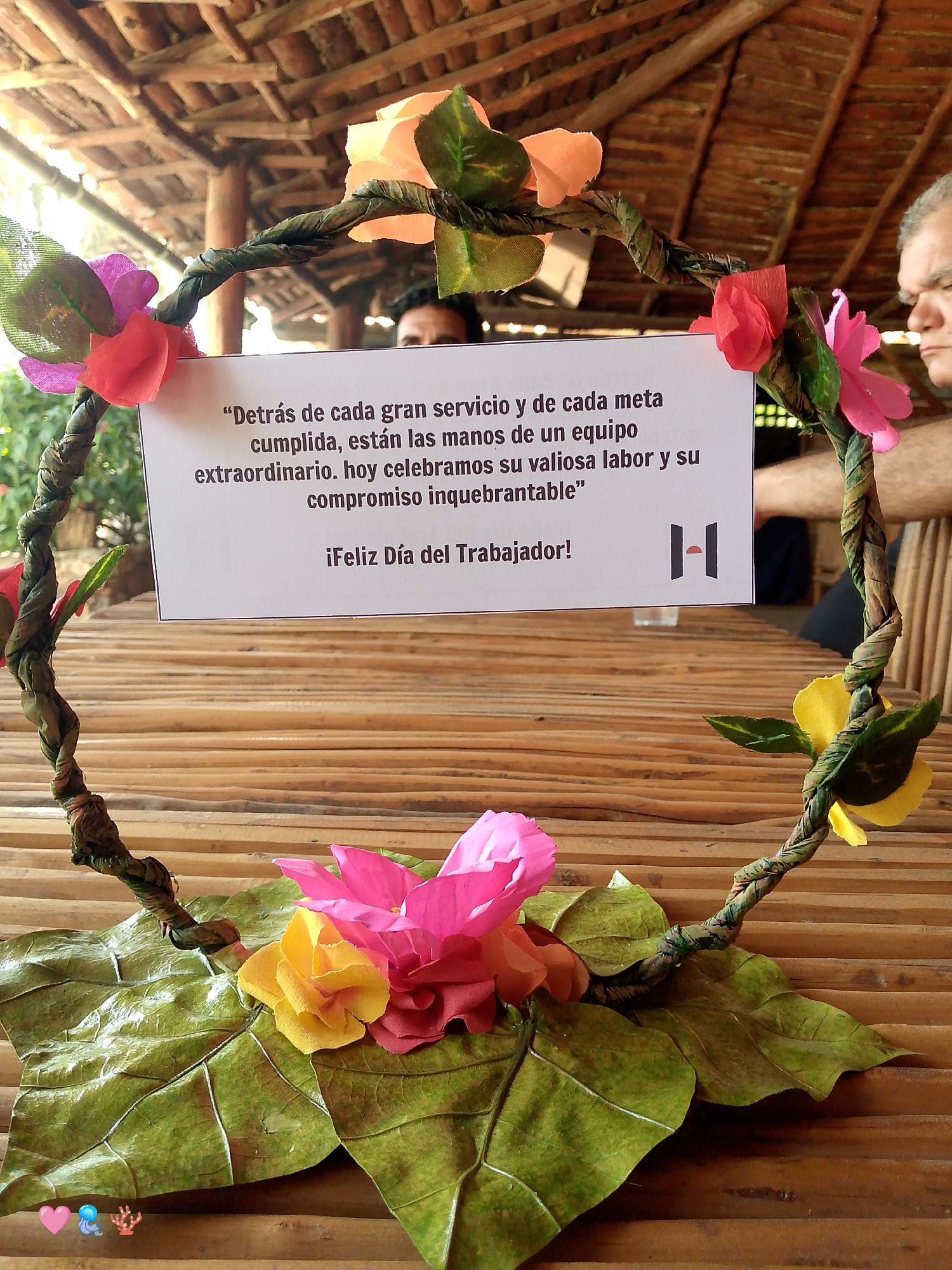 The image captures a close-up view of a floral wreath resting on a bamboo table. The wreath is decorated with colorful paper flowers and leaves. A white sign within the wreath bears a printed message in Spanish, "Detrás de cada gran servicio y de cada meta cumplida, están las manos de un equipo extraordinario. hoy celebramos su valiosa labor y su compromiso inquebrantable" and the words "¡Feliz Día del Trabajador!" beneath. A company logo is on the bottom right of the sign. The bamboo table has horizontal parallel lines. In the background, figures are partially visible, suggesting a restaurant setting. The image appears to be taken in an indoor space in Juan Griego, Venezuela.