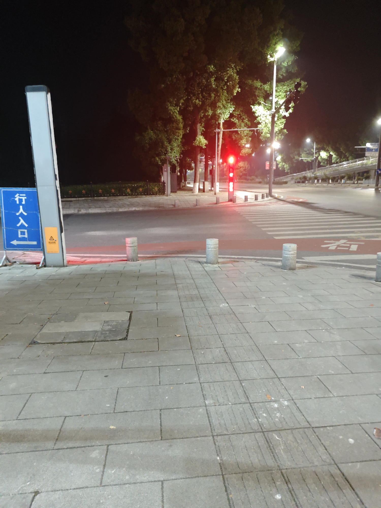 The image shows a night scene at a pedestrian crossing in Chongqing High-tech Zone, China. The foreground consists of a gray tiled sidewalk with tactile paving strips. A blue and white sign with Chinese characters is visible on the left, along with a yellow warning sign.
The pedestrian crossing features a crosswalk with white stripes and a red lane with a white character written on it. Several cylindrical bollards are positioned along the edge of the sidewalk and within the crossing area. A red traffic light is active.
Beyond the crossing is a street with sparse traffic. Trees line the street, lit by streetlights. A bridge or elevated structure is visible in the distance. The sky is dark.