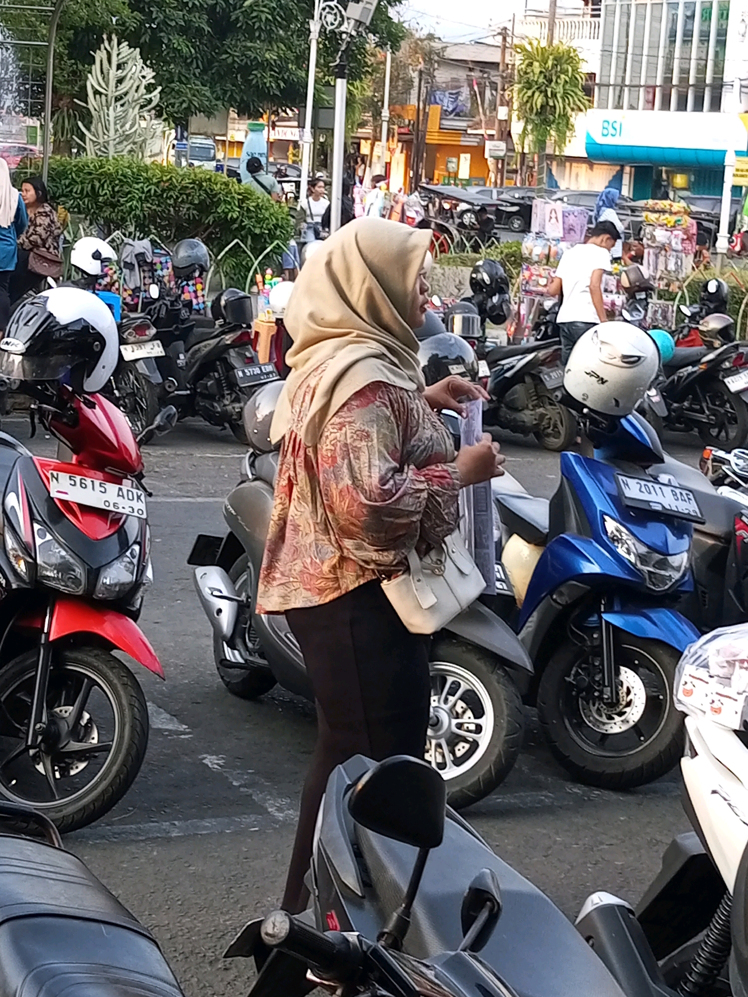 In the city of Batu, Indonesia, this image captures a street scene featuring several parked motorcycles, predominantly scooters. A woman wearing a tan hijab and a patterned shirt stands in the foreground, her hands engaged in holding a small, clear object. Behind her, other motorcycles are visible, some with helmets placed on them. One red scooter in the mid-ground displays the license plate "N 5615 ADK 06-30". Further back, a blue scooter with the license plate "N 2011 BAF" is parked, and a person in a white shirt is seen interacting with a market stall. The environment suggests a commercial or public area with a backdrop of buildings and trees, consistent with a bustling urban setting.
