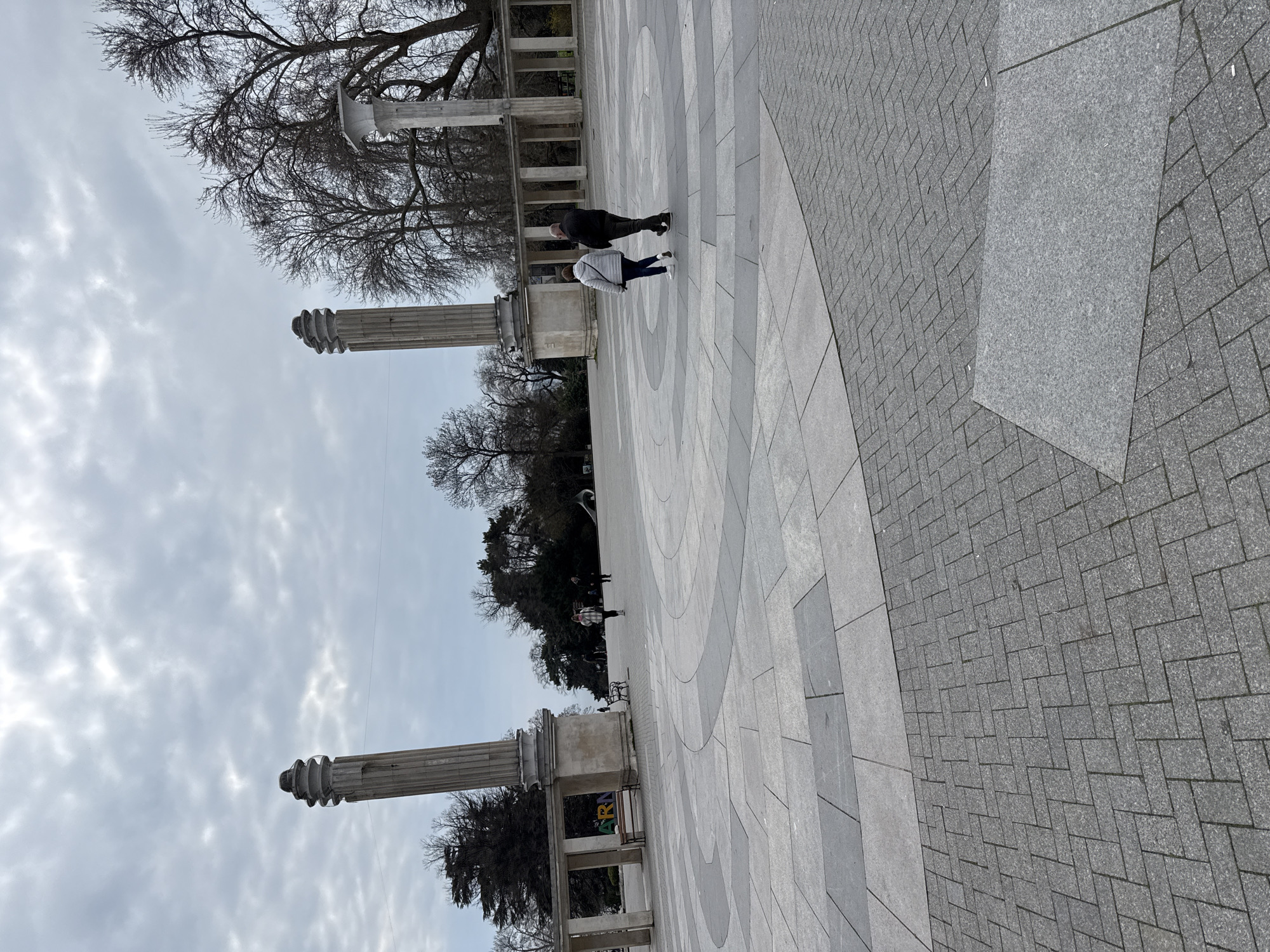 The image depicts a public square in Varna, Bulgaria, characterized by gray stone paving in concentric patterns. The scene includes neoclassical pillars supporting elevated walkways, bare trees, and cloudy sky. Two people are walking in the square. A strip of brick-like paving runs along the right edge of the frame.