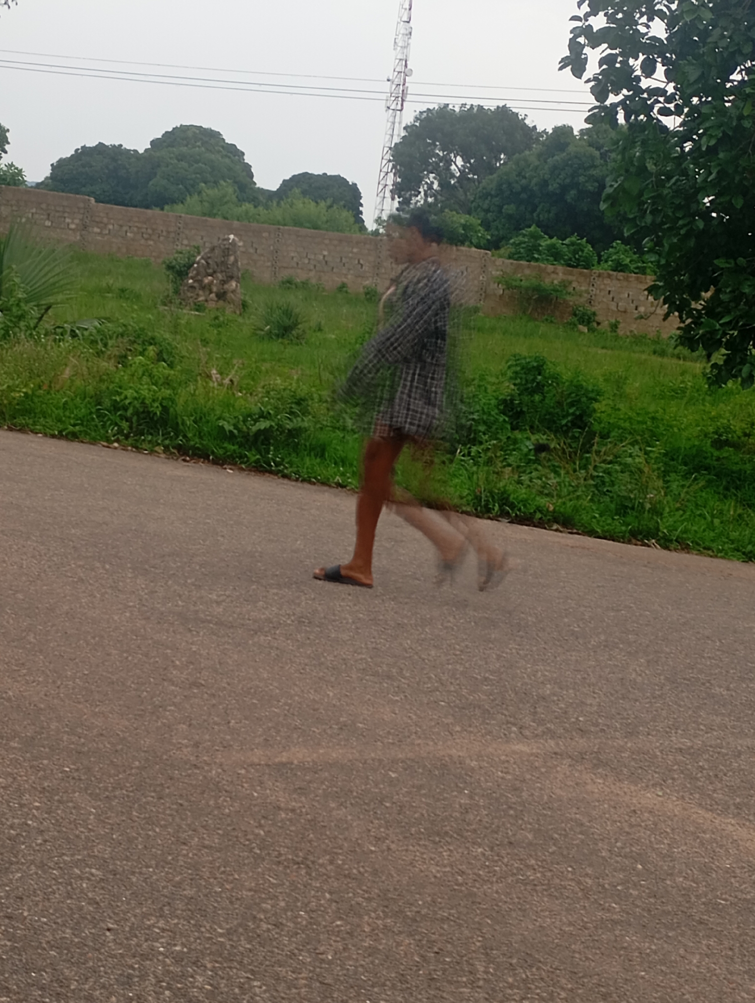 A person is captured in motion, appearing blurred, as they walk across an asphalt road. The individual is wearing a checkered shirt and dark shorts. They have on sandals. The background consists of a low, brick wall, beyond which is lush green vegetation and trees. A telecommunications tower is visible in the distance, along with power lines stretching across the sky. The sky is overcast. The scene appears to be outdoors, on a road in Jos, Nigeria.
