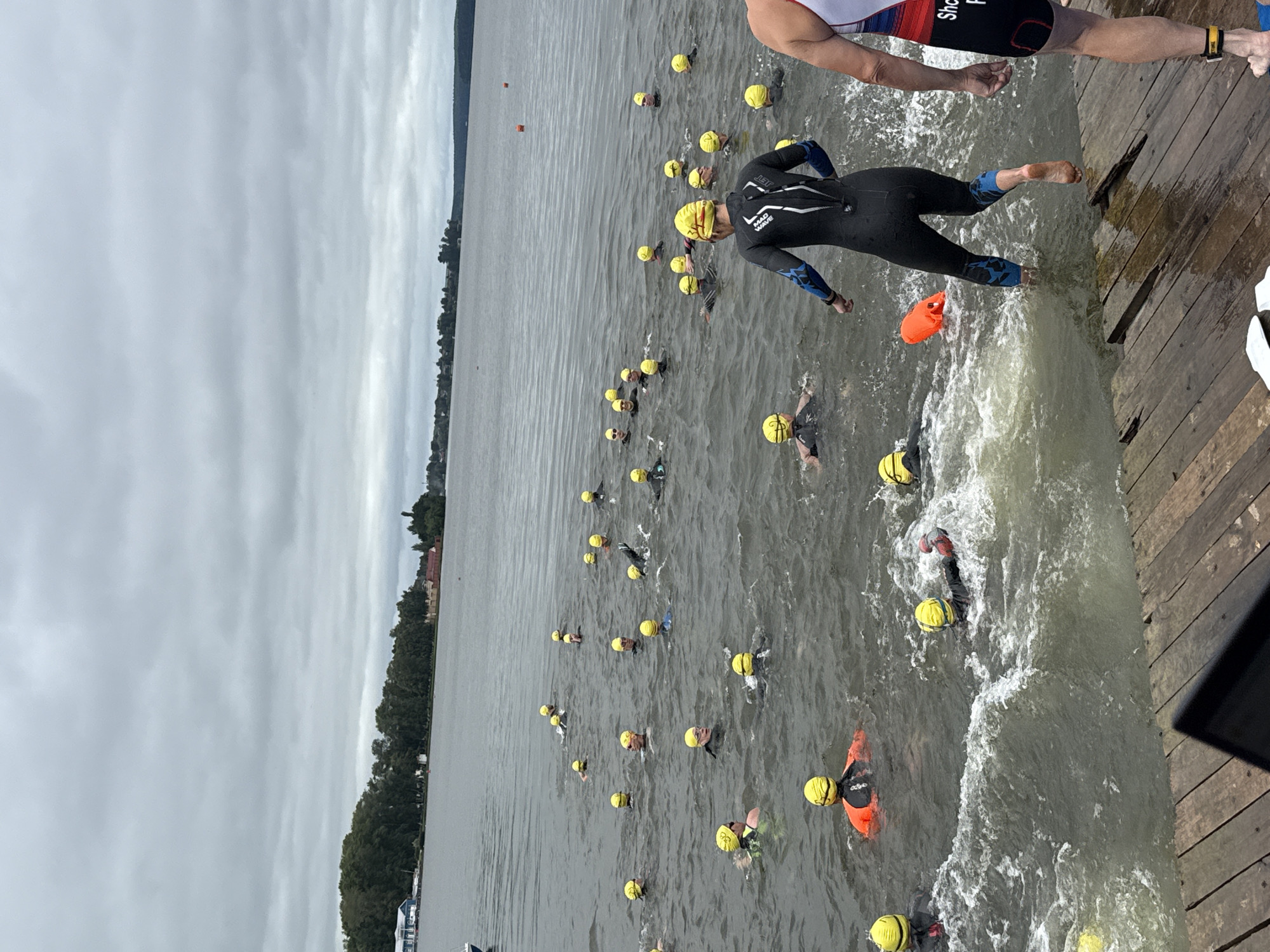 This image depicts a group of swimmers in a body of water, likely during a race or training event, based on the yellow swim caps worn by most participants. The swimmers are dispersed across the water, with some closer to a wooden dock in the foreground and others further out towards the horizon. The water appears slightly choppy, suggesting some wind or activity. In the background, a tree-lined shore is visible, with buildings suggesting a settlement. The sky is overcast with clouds. On the dock in the foreground, the legs of two individuals are visible. One person is wearing a black wetsuit with blue accents, and the other is wearing a white, red, and blue striped athletic top and black shorts. An orange buoy is floating in the water to the left of the person in the black wetsuit. This scene is consistent with an open water swimming event taking place on the Votka River in the city of Votkinsk, Russia, a location known for its aquatic activities.