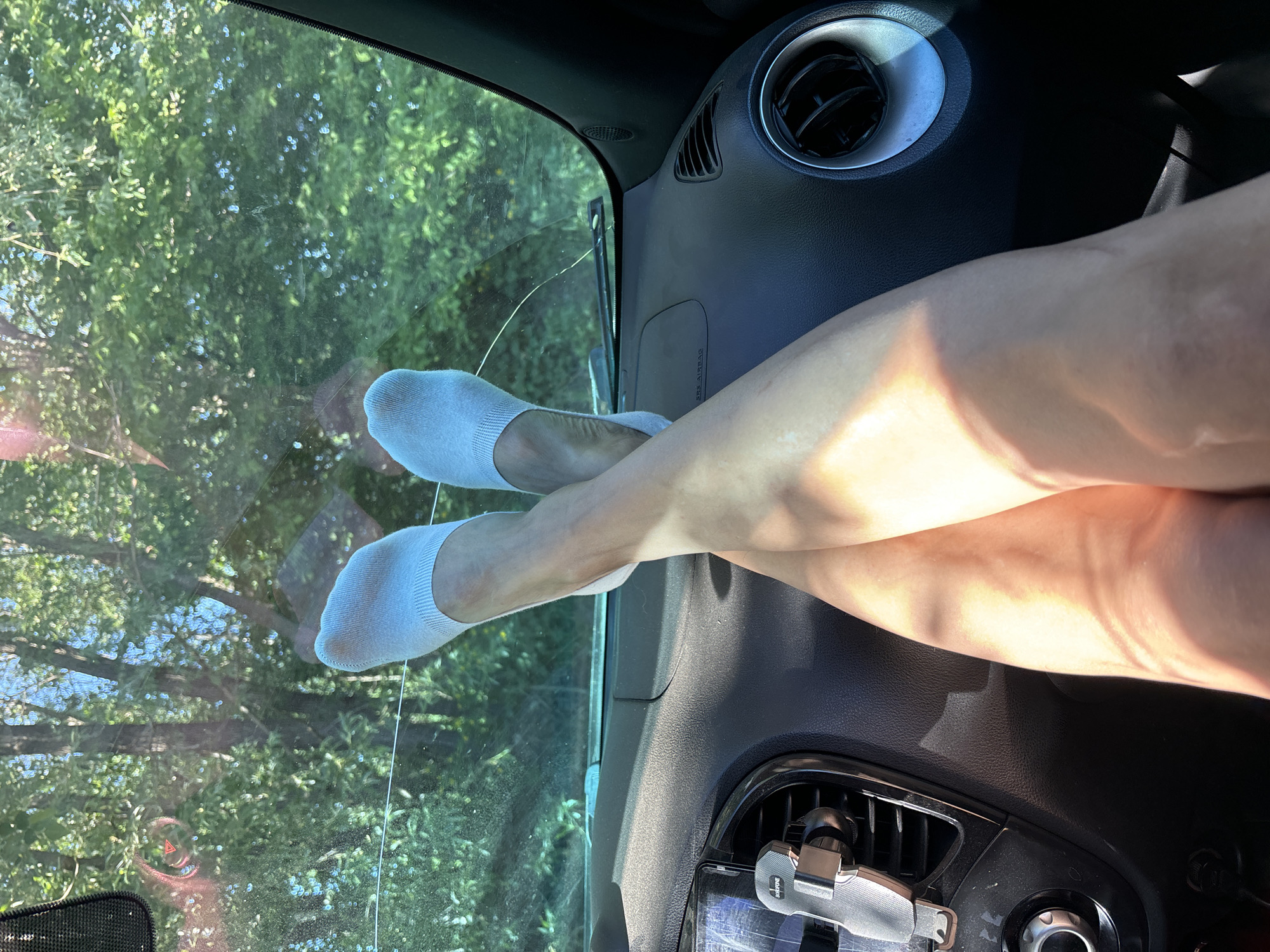 A photograph taken from inside a car shows a pair of legs with white socks resting on the car's dashboard, with feet towards the windshield. The windshield looks out to green trees. The car's dashboard is visible, including air vents. Due to the limited visual information, it's not possible to confirm the specific location in Chaikovsky, Russia.