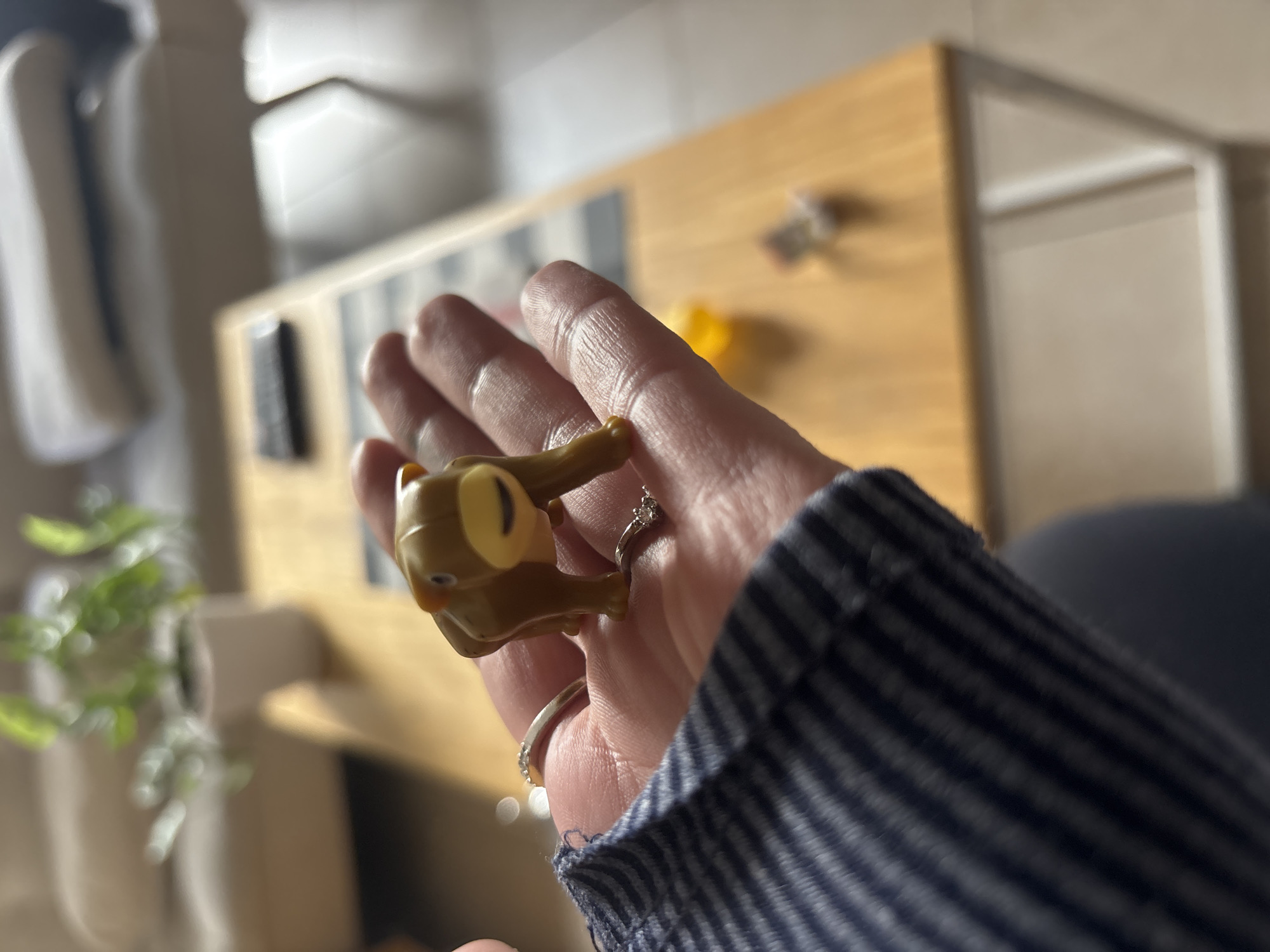 A person's hand holds a brown, cartoonish-looking turtle figure between the fingers. The hand is wearing a blue and white striped sleeve and two rings. The background is blurred, but includes a light brown wooden surface, a plant, and a white object. The image was taken indoors. Location information is unavailable, so cannot be determined if the image was taken in Buenos Aires, Argentina.