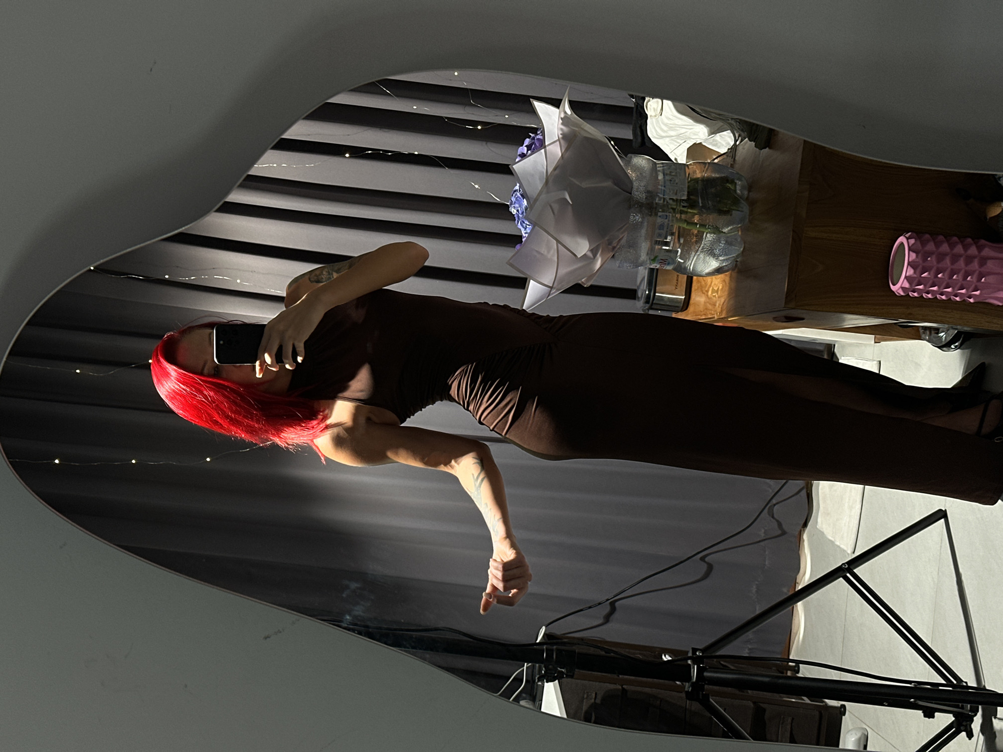 A woman with bright red hair is reflected in a mirror. She wears a brown dress and holds a phone up to take a photo of herself. The mirror's reflection also shows a floral arrangement in a vase, a pink textured cylinder, and what appears to be the frame of a backdrop stand or lighting equipment. The background appears to be a textured surface, potentially a curtain or wall, with string lights. The location, Dalung, Indonesia, is not discernible from the image.