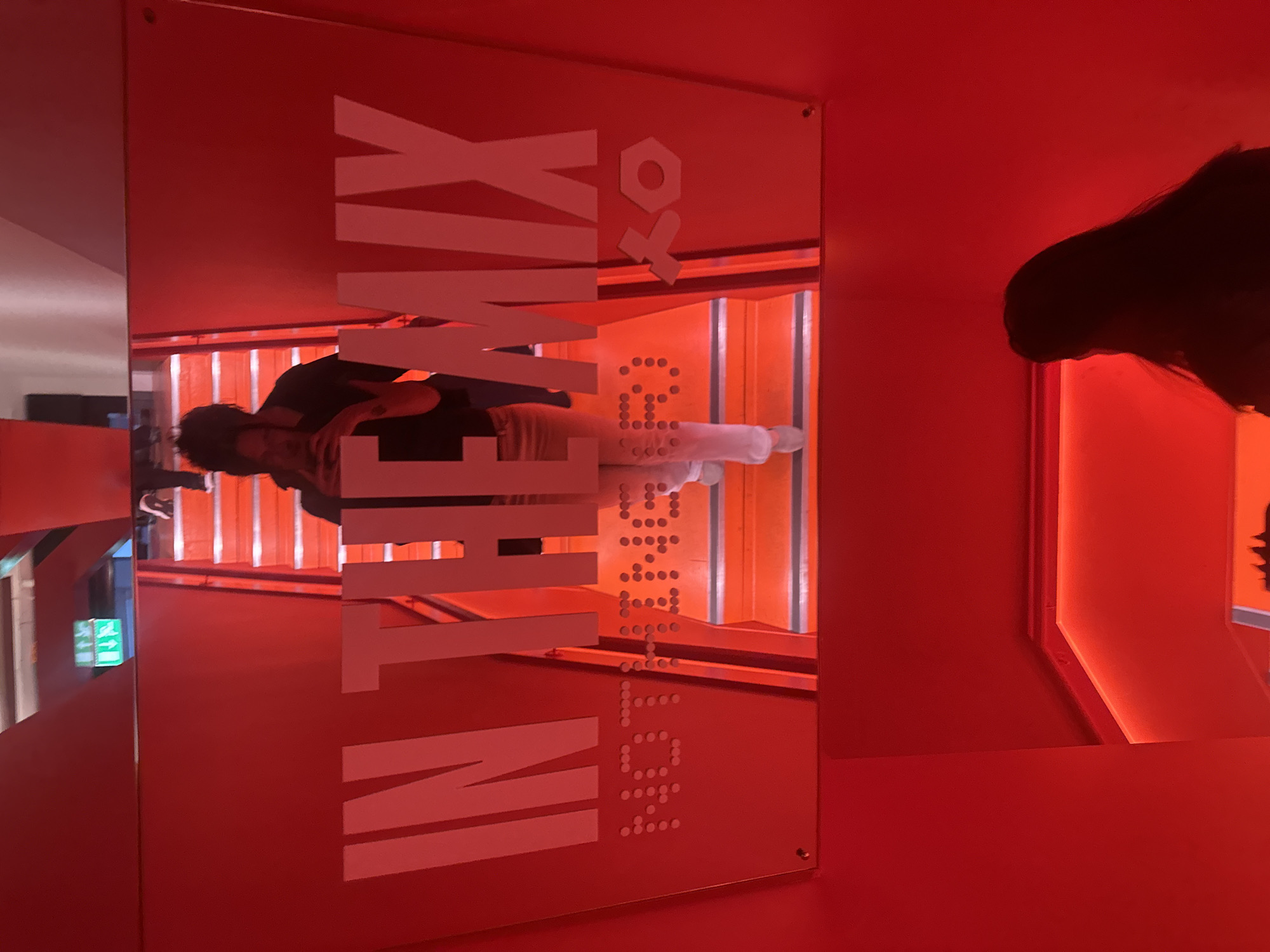 The photograph captures a scene within what appears to be an interior space in London, United Kingdom. The color scheme of the room is predominantly red, with a reflective acrylic or glass panel displaying the words "IN THE MIX KO." The reflection in the panel reveals a set of stairs. Two individuals are partially visible in the reflection. The woman on the right appears to have dark hair and is facing towards the other side of the room, and the man is standing on the stairs. The lighting in the room is artificial. The reflection of an emergency exit sign is on the left side of the panel. The words “IN THE MIX KO” are arranged vertically, with “IN THE MIX” spelled out in solid white letters and “KO” rendered in white.