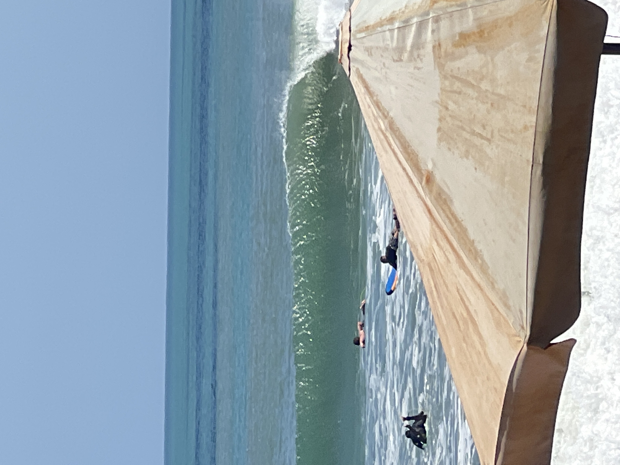 A beach umbrella with a light brown canopy dominates the foreground of this eye-level, outdoor, vertical, medium shot of Kuta beach, Indonesia. The umbrella is casting a shadow on the white frothy waves breaking on the shore. Beyond the umbrella, four people are visible in the turquoise ocean. One person in a black wetsuit is partially submerged in the water, with their arms raised. To their right, two people are lying on surfboards. One is on a blue and orange board, while the other is on a white board. Further out, another person is lying on a white surfboard, paddling in the water. A large, green wave is breaking in the midground, with white foam cresting. The horizon line separates the ocean from a clear, pale blue sky.