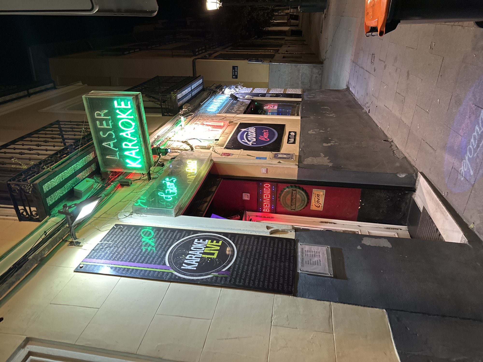 The image is of a building in Madrid, Spain, featuring the karaoke bar "Laser Karaoke Cheers" with signs in English and Spanish. A large black sign with the words "Karaoke Live" in a stylized font is affixed to the wall. The entrance is adorned with neon signs, including "Cheers" and "Karaoke Cheers." An "Open" sign and a "Bienvenido" sign are also visible. Other signs advertise Karaoke in different music styles. The street outside appears to be a narrow pedestrian zone, with the street name "Calle de San José" displayed. A blurred image on the sidewalk reads "Karaoke." The lighting suggests it is night, with illuminated signs highlighting the location.