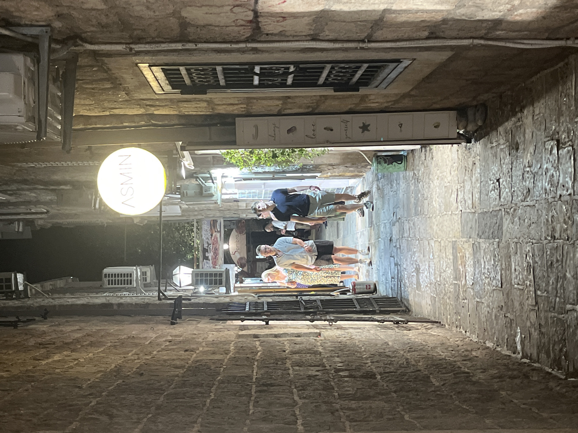 In the city of Budva, Montenegro, a narrow, stone-paved alleyway is illuminated by overhead lights and a circular sign displaying "ASMIN" in red lettering. Two men and a woman are walking down the alley. The man in the foreground on the right wears a dark blue polo shirt and gray shorts, while the man beside him, to the left, is in a light blue collared shirt and dark shorts. The woman in front of them wears a floral print dress. Shops line the alley walls, some with open doorways and illuminated interiors. The stone walls of the buildings are aged and textured. A decorative sign with the words "always love yourself" and star and heart motifs is mounted on the wall to the right of the alley. The overall scene suggests a bustling evening in an old town setting.