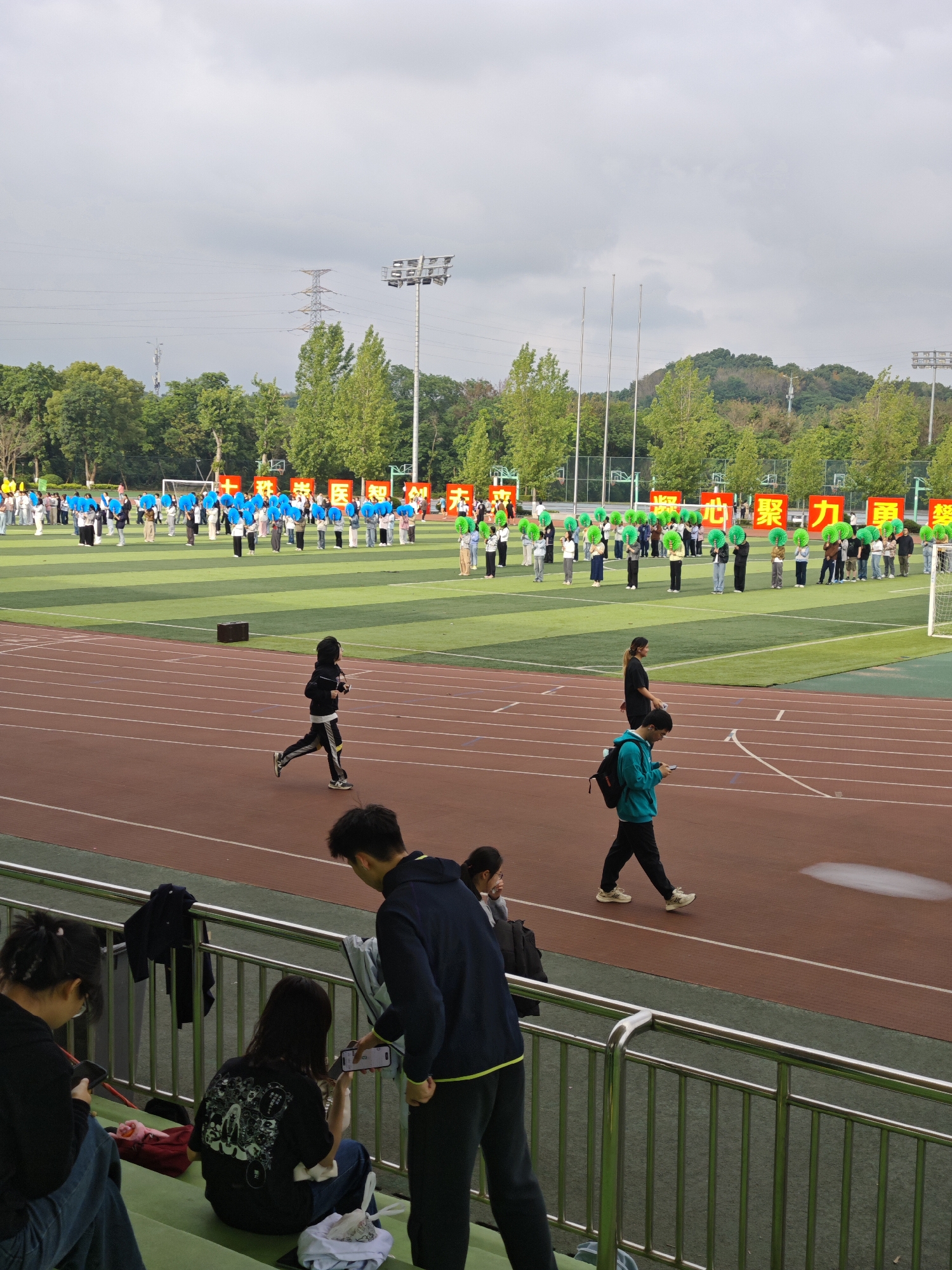 This outdoor photograph, taken in the Chongqing High-tech Zone, China, captures a scene at a sports stadium under an overcast, cloudy sky.
The main field, covered in green artificial turf, is filled with a large group of people arranged in orderly rows. They are participating in an organized event, with many holding either vibrant blue or bright green fan-like props, forming distinct sections of color. Behind these participants, several large red signs display Chinese characters. Visible characters include "学医" (xué yī - study medicine), "智创" (zhì chuàng - intelligent creation/innovation), "聚力" (jù lì - gather strength), and "勇" (yǒng - brave/courage), suggesting a slogan or motto related to education or a specific institution, possibly medical. In the background, lush green trees, distant hills, utility poles, and tall stadium lights complete the setting.
On the red running track surrounding the field, a person dressed in black and yellow is actively running. Further along the track, another individual wearing a teal hoodie and black pants walks casually, looking at a mobile phone.
In the immediate foreground, from the vantage point of green bleachers, several people are observed. Two young women are seated, both engrossed in their mobile phones. A young man stands beside them, also interacting with a phone held by one of the seated women. Another person is partially visible on the far left. The individuals in the bleachers appear relaxed, possibly spectating or taking a break from the event unfolding on the field. The scene depicts a daytime community or institutional event, characterized by organized activity on the field and casual observation from the sidelines.