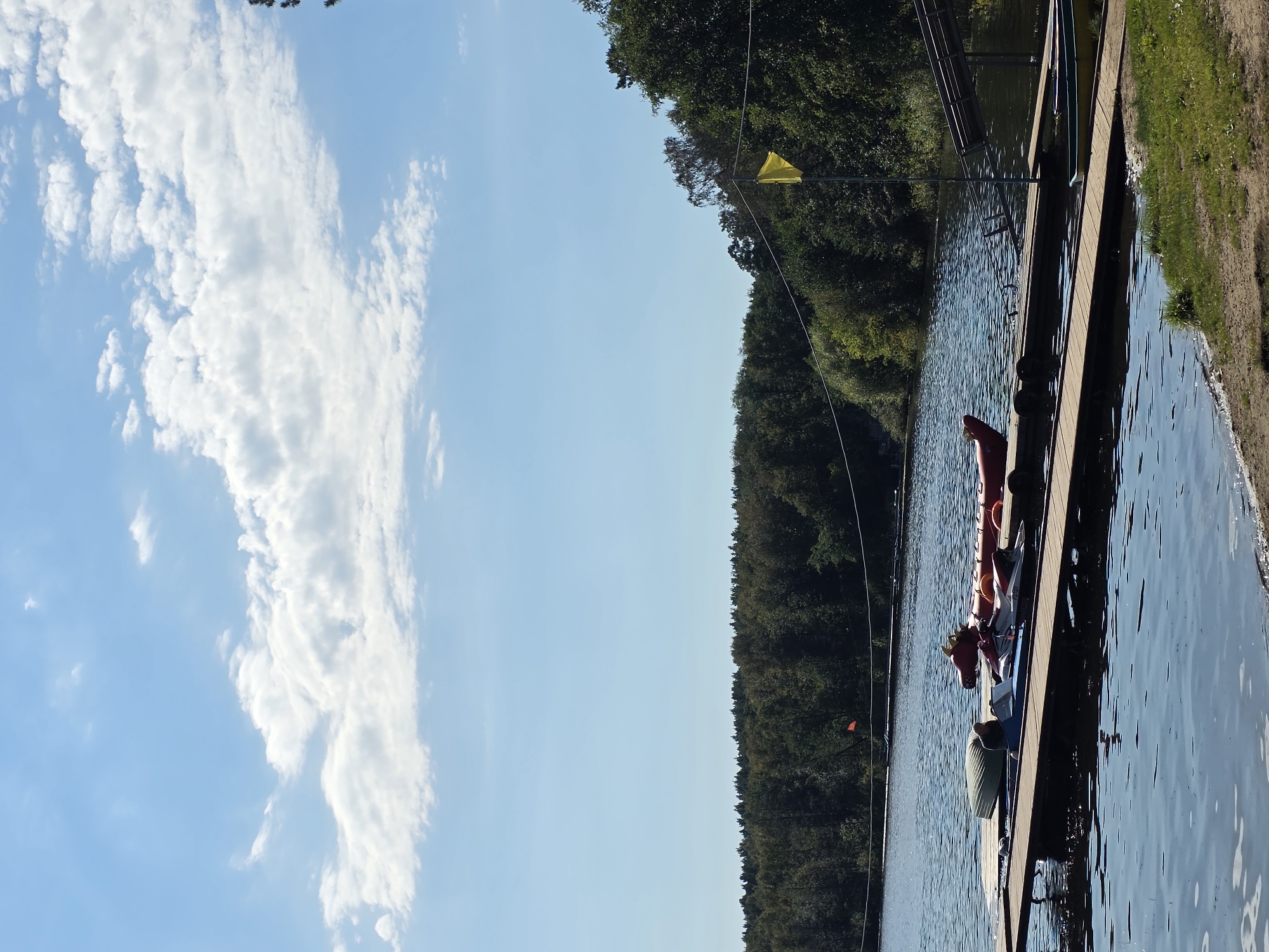 The image shows a wide, calm lake bordered by a dense, dark green forest. A wooden pier extends into the water, equipped with several watercraft, including a red inflatable dragon-shaped boat, a jet ski, and a green rowboat. A person is seated in the blue jet ski. A yellow flag is planted on a pole to the right of the pier, near the edge of the forest. The sky is a light blue with scattered white clouds. The foreground features a sandy beach and green grass. This scene is consistent with a recreational area on the shore of Lake Chebarkul in Chebarkul, Russia.