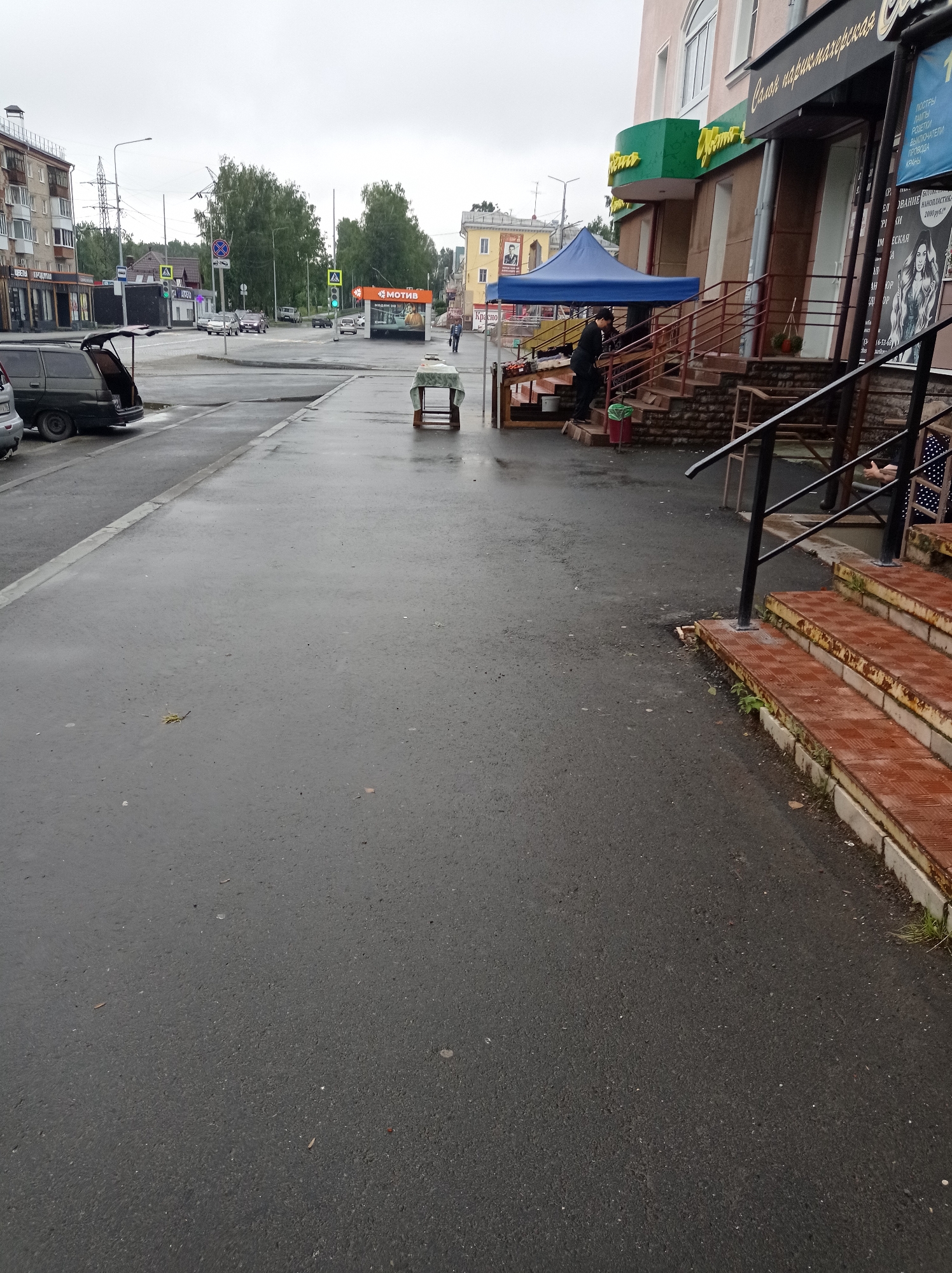 The image depicts a street scene in Degtyarsk, Russia. On the left, a dark-colored minivan with its trunk open is parked on the asphalt. The street is wet, suggesting recent rain, with puddles and scattered leaves. To the right of the minivan, a sidewalk runs along a building with several storefronts, including one with a green awning and Cyrillic signage. A blue canopy is set up on the sidewalk, and a person in dark clothing is standing near some steps, possibly arranging goods. Adjacent to the building, a set of outdoor stairs with rusty metal railings leads upwards. Further down the street, traffic lights are visible, and other vehicles are present. The overall atmosphere is overcast.