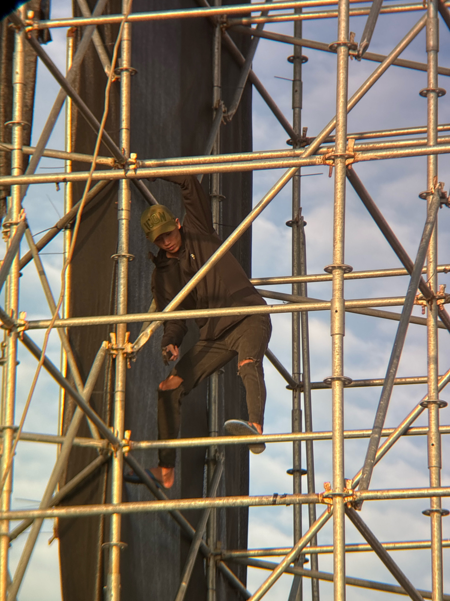 An individual is observed climbing on scaffolding, possibly for event setup or maintenance, in Nha Trang, Vietnam. The scaffolding is constructed from metal poles and connectors, forming a complex, multi-level structure. The person is wearing a dark jacket, dark pants with rips on the knee, a baseball cap with "ICON" written on it, and sandals. The individual's body is positioned in a climbing motion, with one hand gripping a horizontal pole and the other reaching upwards. Their legs are bent, and one foot is visible on a horizontal support. Behind the scaffolding, a dark fabric or banner is partially visible, as well as a sky with clouds. The lighting suggests it is daytime, with the sun casting warm tones on the metal structure. The scene appears to be outdoors, likely in an urban or event-related setting within Nha Trang.