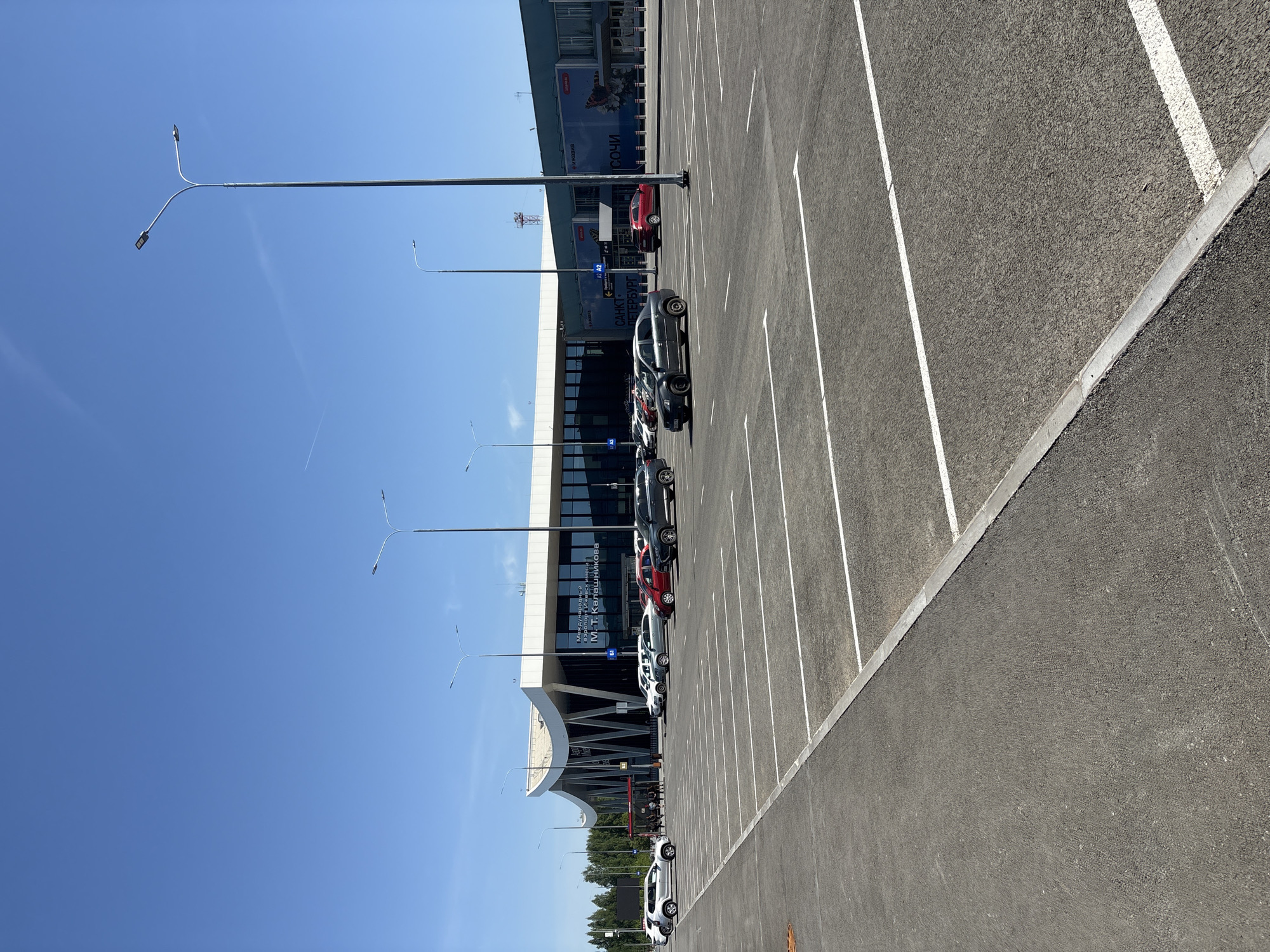 The image captures an outdoor parking area located at an airport in Новое Мартьяново, Russia. The clear blue sky suggests a daytime setting. A line of parked cars, primarily white, gray, and red, occupies the parking spaces adjacent to the airport building. The building has a modern architectural design with glass windows and signage visible on the façade. Streetlights are positioned along the edge of the parking area, extending upwards into the sky. The parking lot is marked with white lines delineating the parking spaces. The surface of the parking area is asphalt.