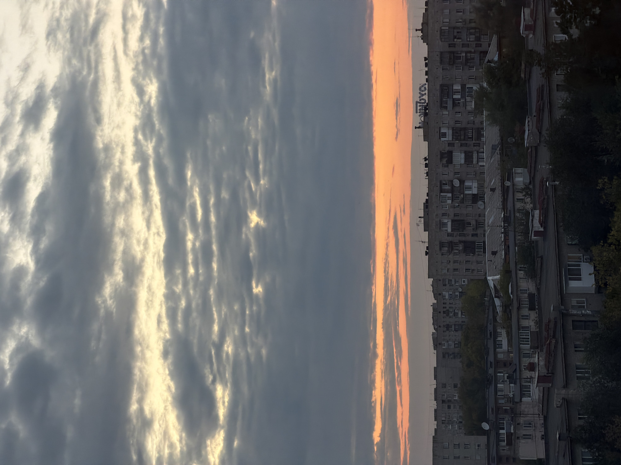 The image depicts a view of an urban residential area at dusk, likely in Pavlodar, Kazakhstan. The sky is filled with a layer of dark, textured clouds that are illuminated from below by a band of orange and yellow light, suggesting the setting sun. Below the sky, a dense arrangement of Soviet-era apartment buildings, characterized by their uniform grey facades and numerous windows, stretches across the frame. The buildings vary in height and architectural style but share a common, utilitarian design. Some structures have satellite dishes visible on their roofs. The foreground shows the roofs of several lower buildings, some with darker, tiled roofs and others with lighter-colored surfaces. Trees and foliage are interspersed between and behind the buildings, particularly in the mid-ground, adding a touch of green to the otherwise grey urban landscape. A prominent sign with Cyrillic lettering, "NOVAYА," is visible on the rooftop of one of the taller apartment buildings in the mid-ground. There are no people visible in the image, and the scene appears to be devoid of any dynamic action, presenting a static, architectural cityscape at twilight.