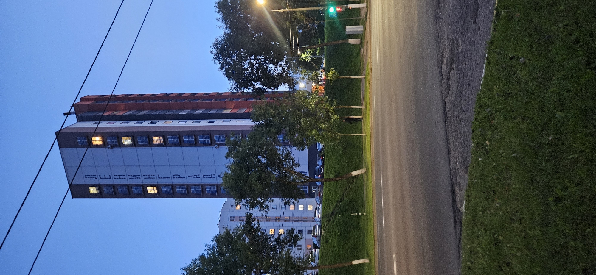 The image shows a multi-story residential building with the Cyrillic letters "ЛЕНИНГРАД" written vertically on its facade. The building is colored white, with red and dark grey sections. Several windows are illuminated from within, suggesting occupancy. In front of the building, a grassy slope is visible, with trees planted along its crest. Below the slope is a paved road with a white line marking the lane. In the background, other residential buildings and parked cars can be seen. A traffic light with a red signal is visible on the right side of the image. The sky is a deep blue, indicating it is either dawn or dusk. The presence of the text "ЛЕНИНГРАД" (Leningrad) on the building strongly suggests this location is in Russia, as it refers to the former name of Saint Petersburg. The information provided in the prompt claiming the location is Vitsebsk, Belarus is incorrect. The scene depicts a typical urban environment with residential architecture and street infrastructure.