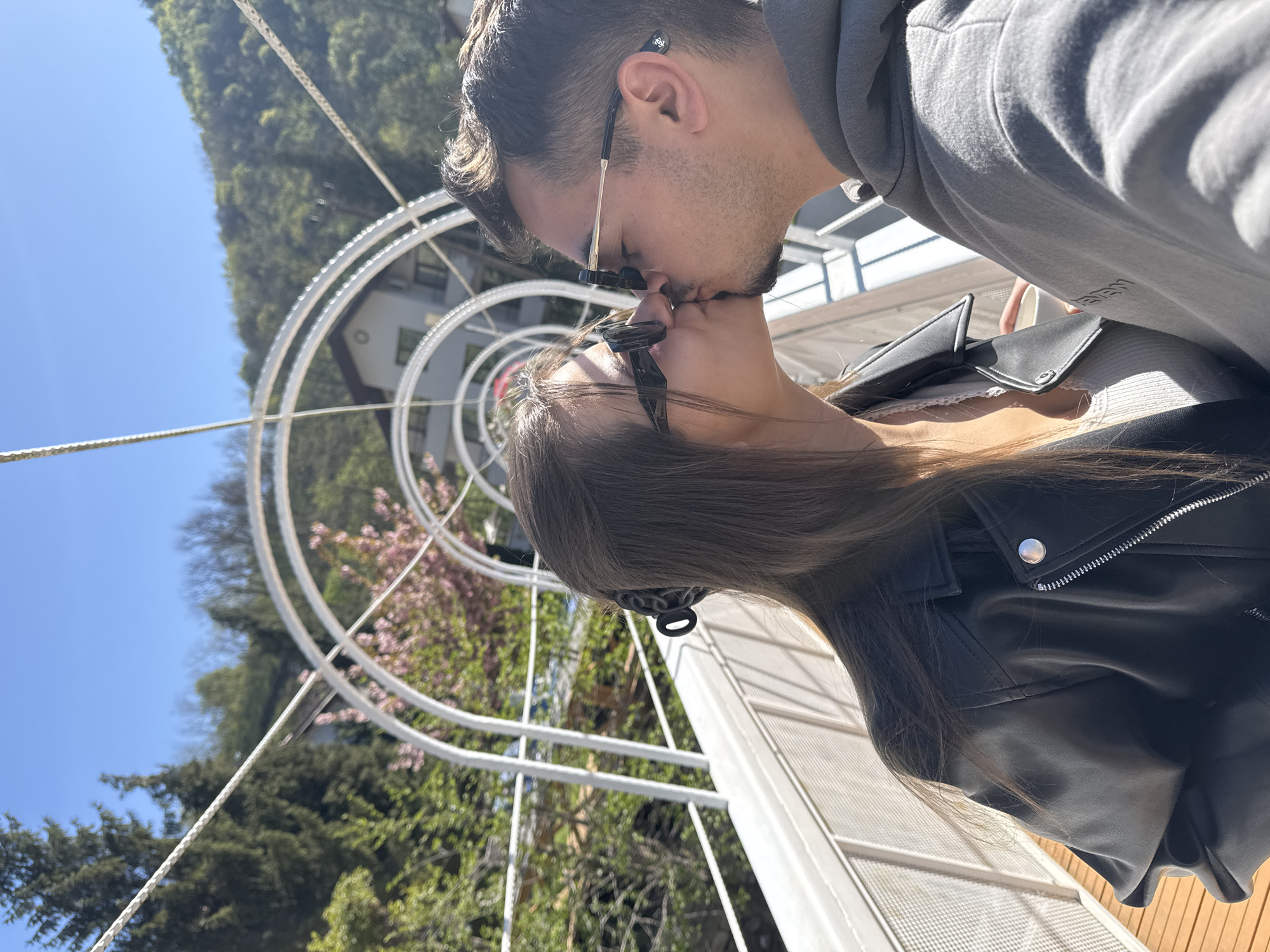 A couple is kissing in front of the St. Mina chapel in Marchevo, Bulgaria. The man is wearing a gray hoodie and sunglasses, while the woman is wearing a black leather jacket and sunglasses. They are standing in front of a white, metal, circular structure. Behind the structure, there are trees and a building. The sky is blue.