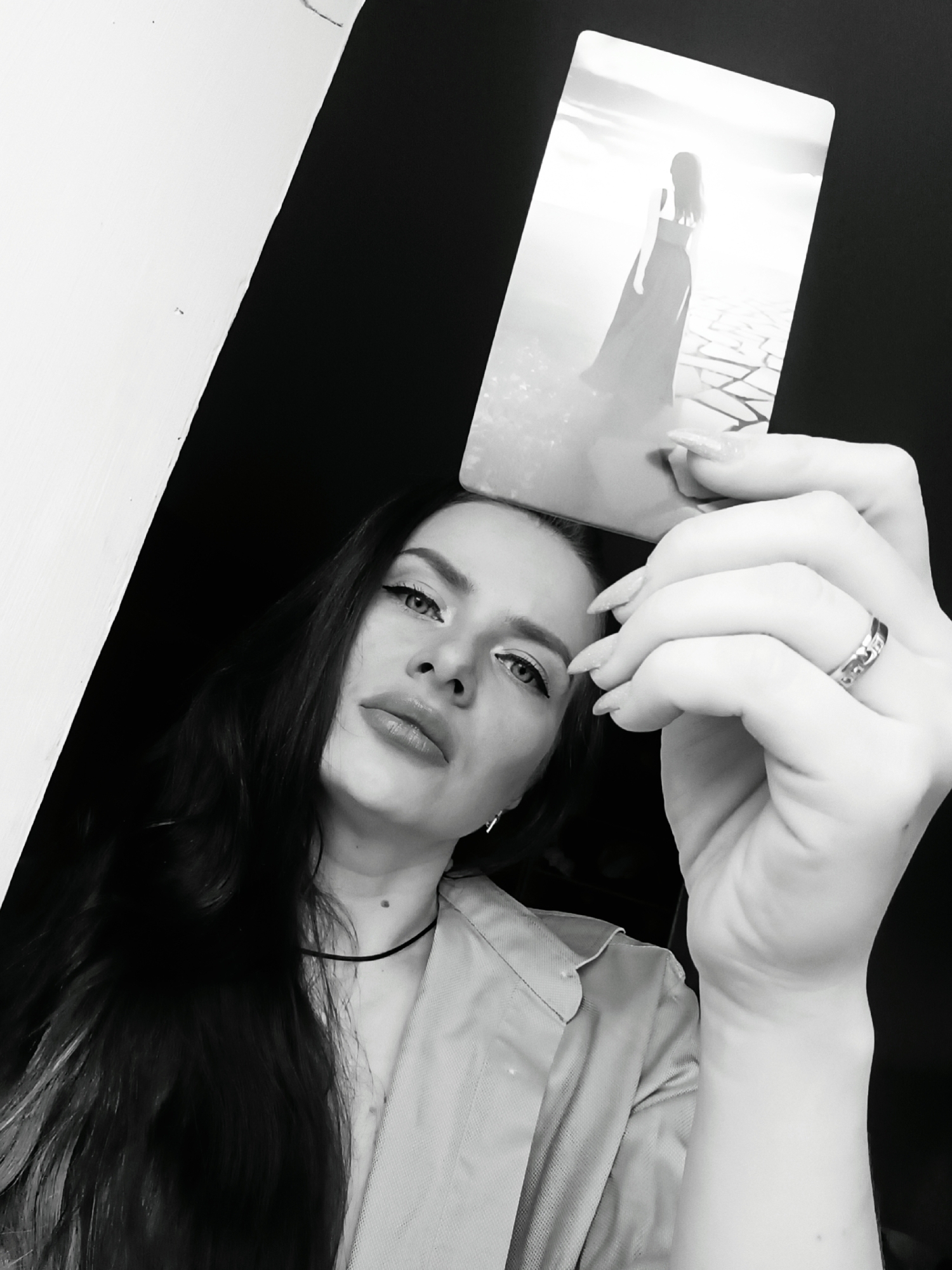 A black and white, low-angle portrait depicts a young woman holding a rectangular card above her head. The woman has long, dark hair, full lips, and distinctive winged eyeliner, looking directly at the viewer with an intense and captivating expression. She is wearing what appears to be a dark choker necklace and a light-colored, collared shirt or jacket.
Her right hand, with long, manicured nails, is prominent in the foreground, holding the card. A ring with faint, unreadable engravings is visible on her ring finger. The card itself features a high-contrast image of a silhouetted woman, seen from the back, with long hair and a flowing dress, gazing towards a bright, almost ethereal background that suggests a sky or distant landscape. The ground beneath the silhouetted figure has a cracked or geometric pattern.
The background of the main image is predominantly dark, creating a strong contrast with a diagonal white element on the left side, possibly a wall or architectural feature. The overall aesthetic is dramatic and somewhat mysterious, emphasized by the monochrome palette. No explicit text is clearly visible on the card or the ring. The setting is indoors, with the location context provided as Rusalevka, Russia.